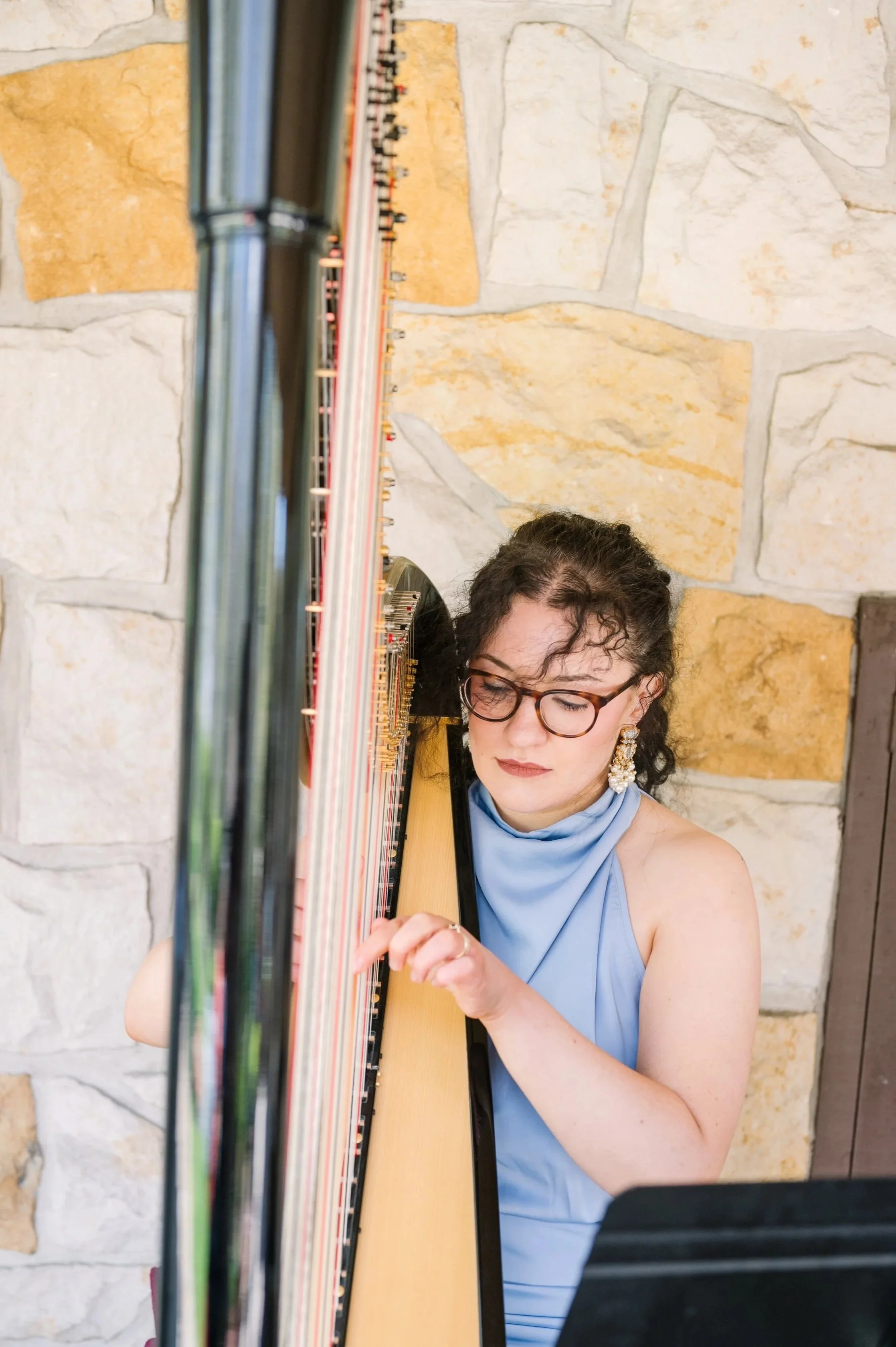 A woman with dark curly hair, glasses, and large floral earrings is playing a harp. She is wearing a light blue sleeveless dress and is sitting in front of a stone wall.