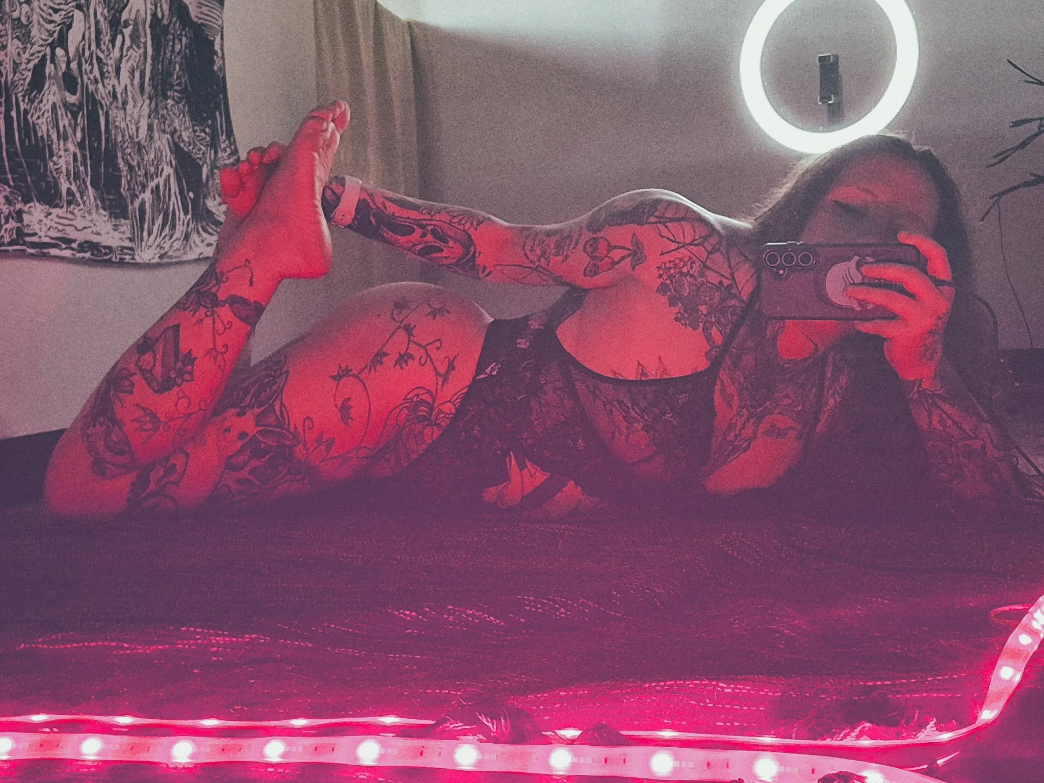 A woman with tattoos on her arms and legs taking a selfie in front of a mirror, with a circular light fixture and a potted plant in the background. The photo has red lighting effects.