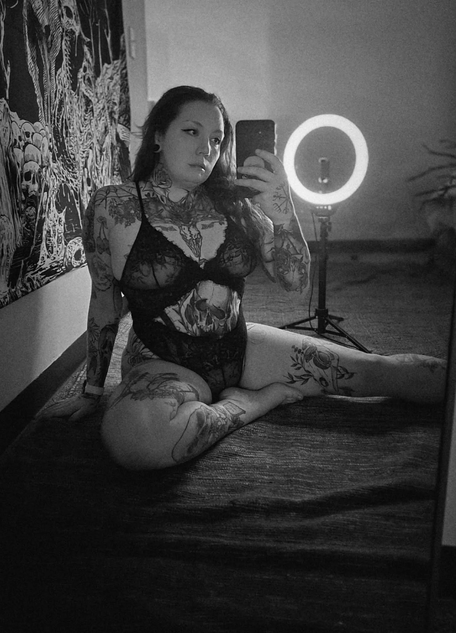 Black and white photo of a woman with tattoos, sitting on a rug, taking a mirror selfie with ring light in the background.