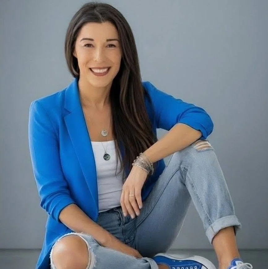 Smiling woman with long dark hair wearing a blue blazer, white t-shirt, ripped jeans, and blue sneakers, sitting on the floor against a neutral background.
