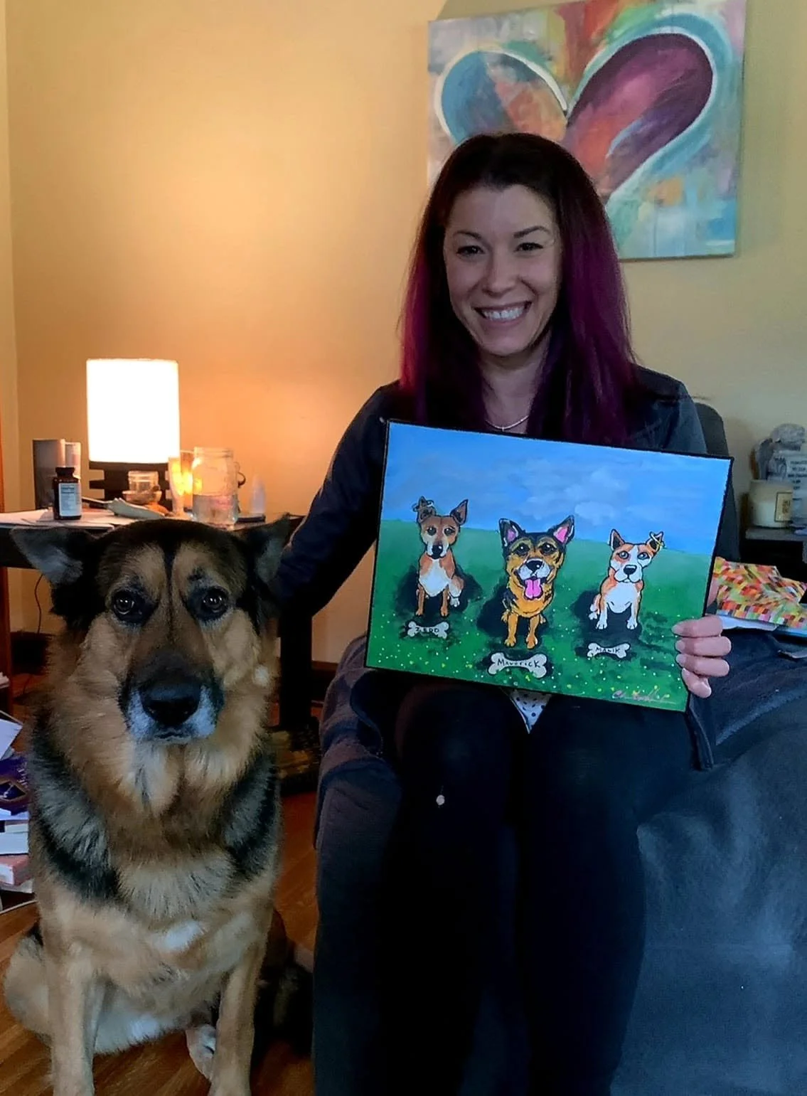 A woman with red and purple hair holding a colorful painting of three dogs, sitting on a black chair in a living room. A large dog is sitting beside her. The background features a wall with a colorful heart painting, a table with lamps, bottles, and papers.