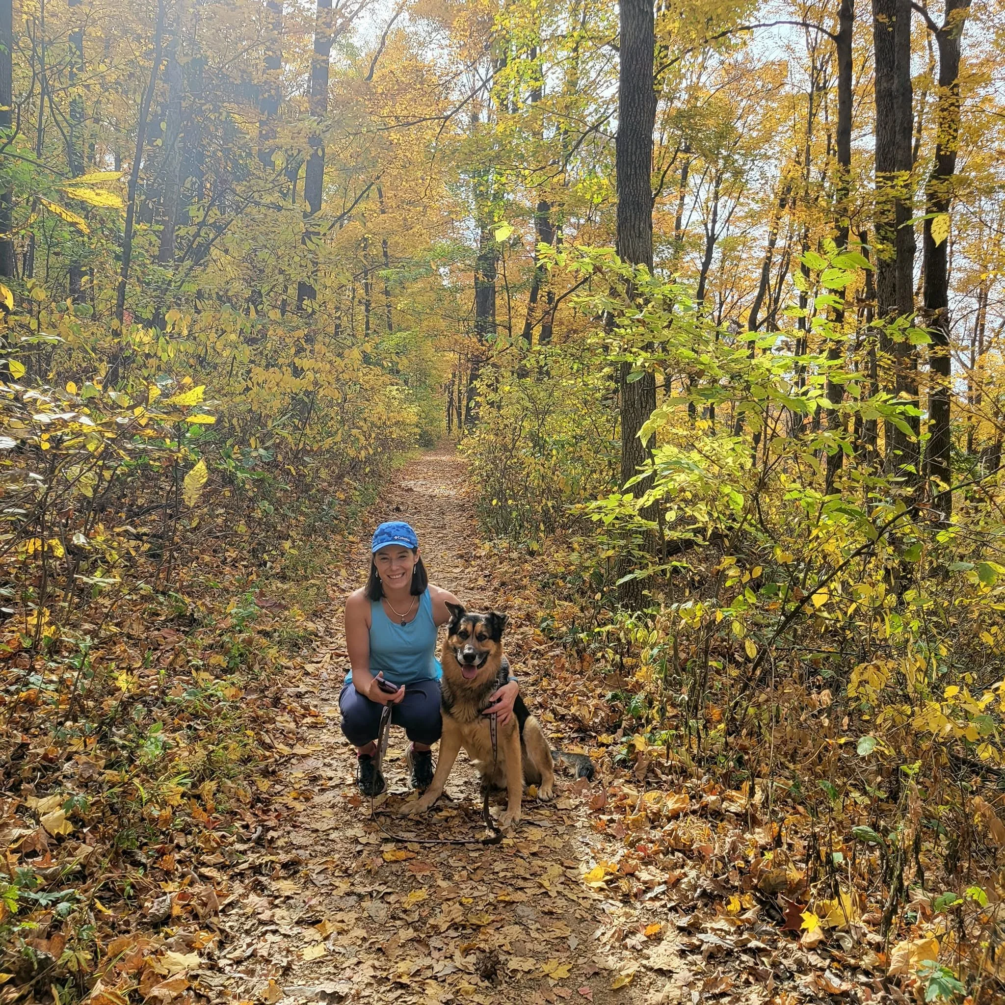 Woman with a dog on a leash, smiling, kneeling on a leaf-covered trail in a forest during autumn, with trees with yellow and orange leaves.