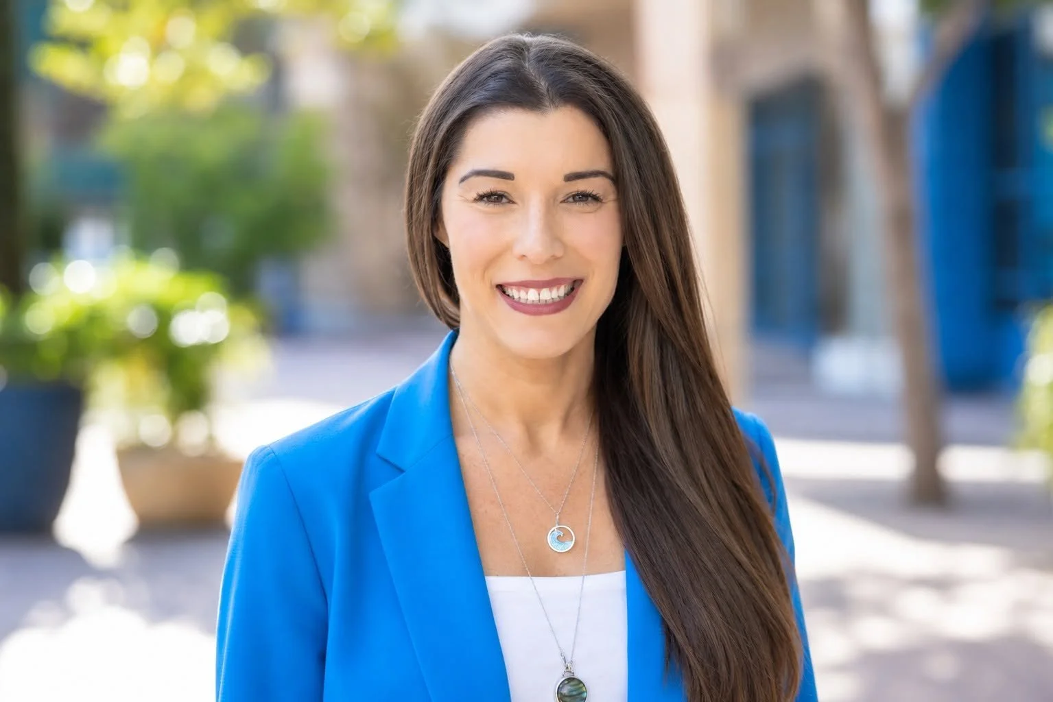 A woman smiling outdoors, wearing a blue blazer and layered necklaces, with trees and a blue mail booth in the background.