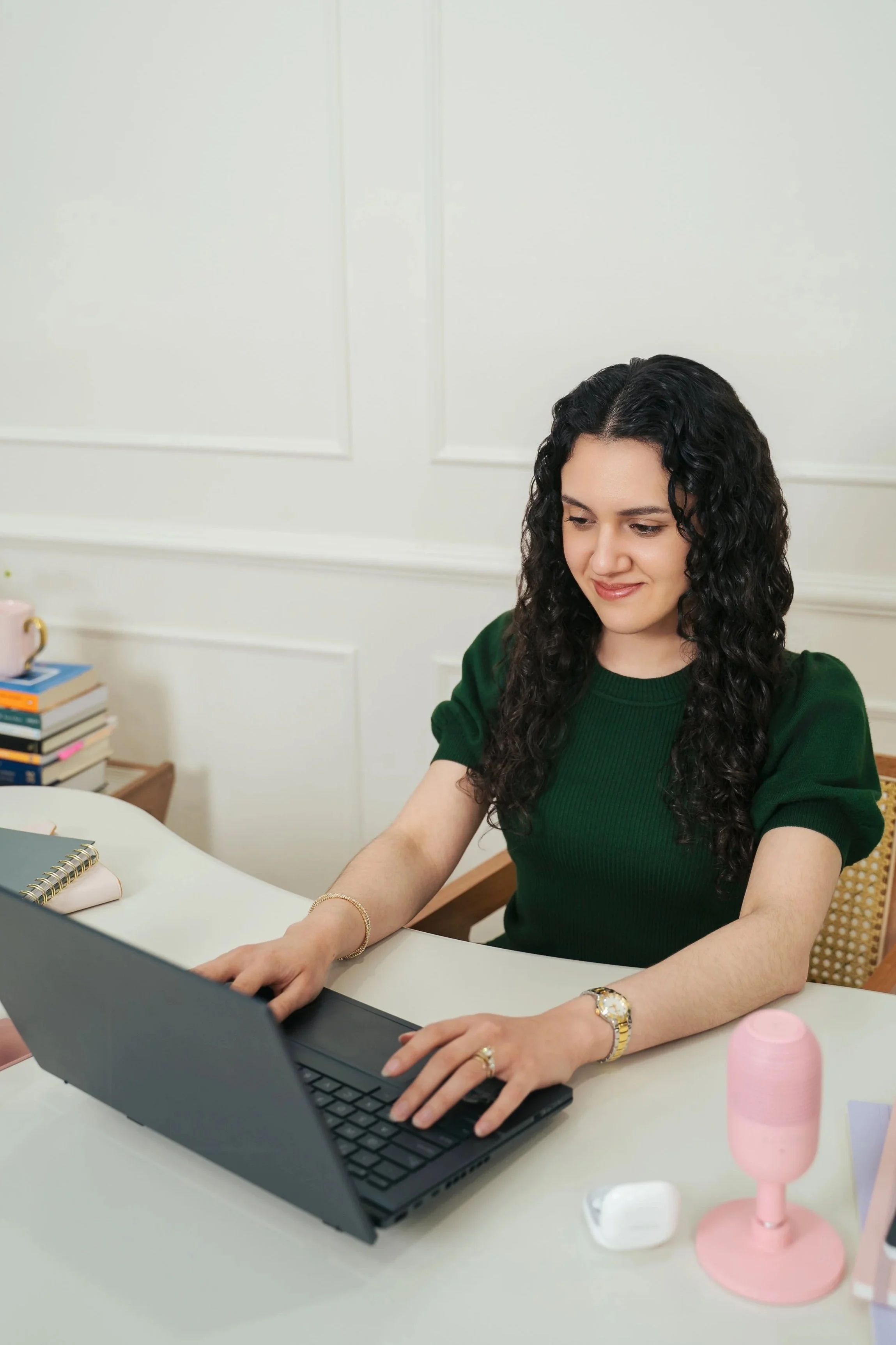 Woman with curly dark hair sitting at a white table, using a black laptop. She wears a green top, gold watch, and rings. Behind her is a white wall with decorative moldings, and to her left are stacked books, a pink mug, and a pink lamp.