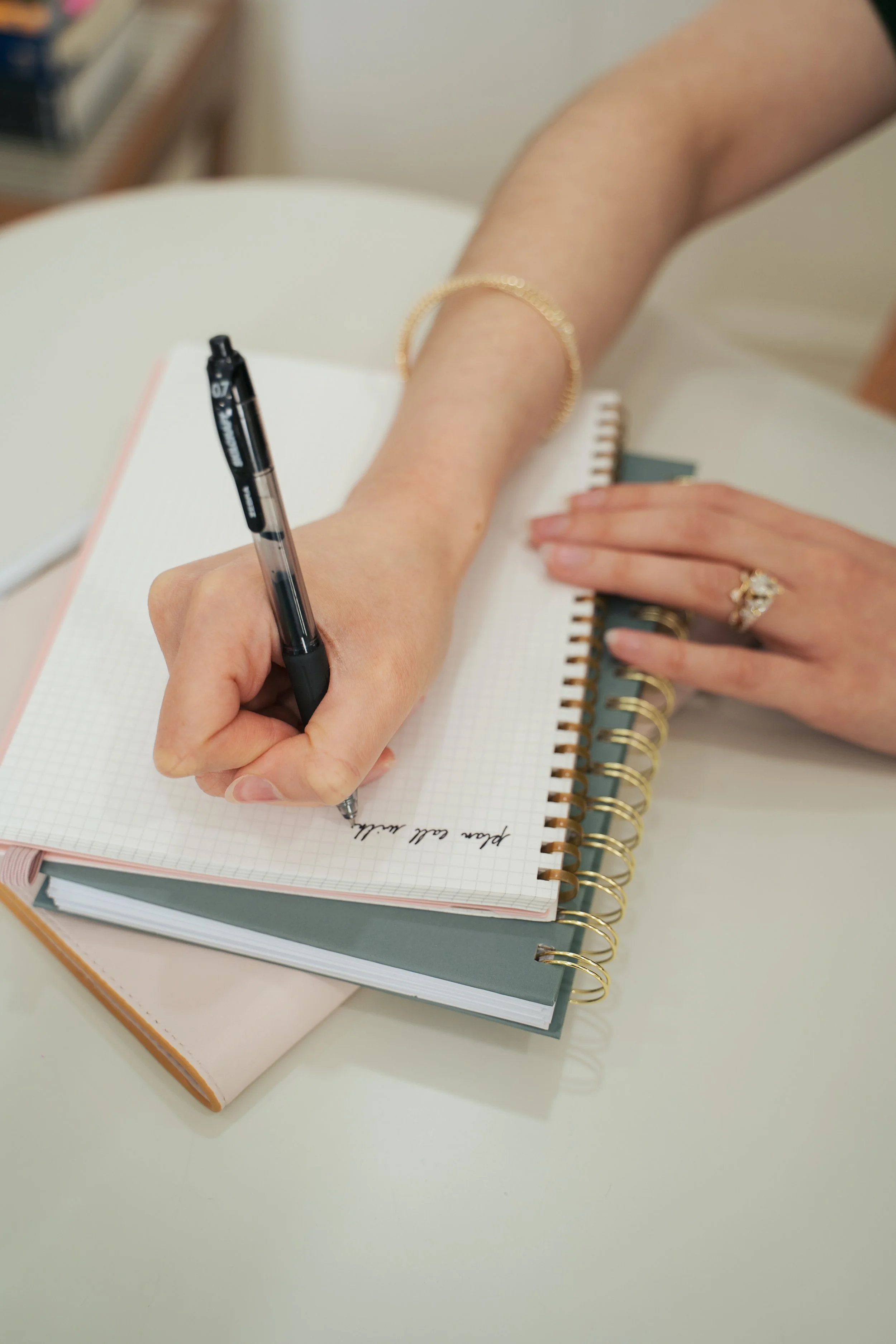 A person writing in a notebook with a black pen, wearing jewelry on their wrist and fingers, with multiple notebooks stacked on a white table.