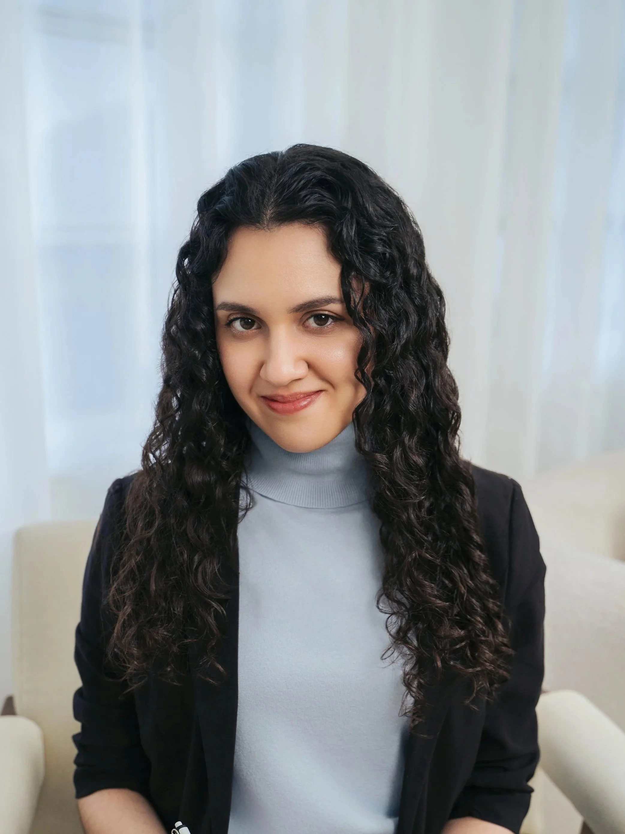 A woman with long curly black hair wearing a light gray turtleneck and a black blazer, smiling softly, seated in a bright indoor setting with white curtains in the background.