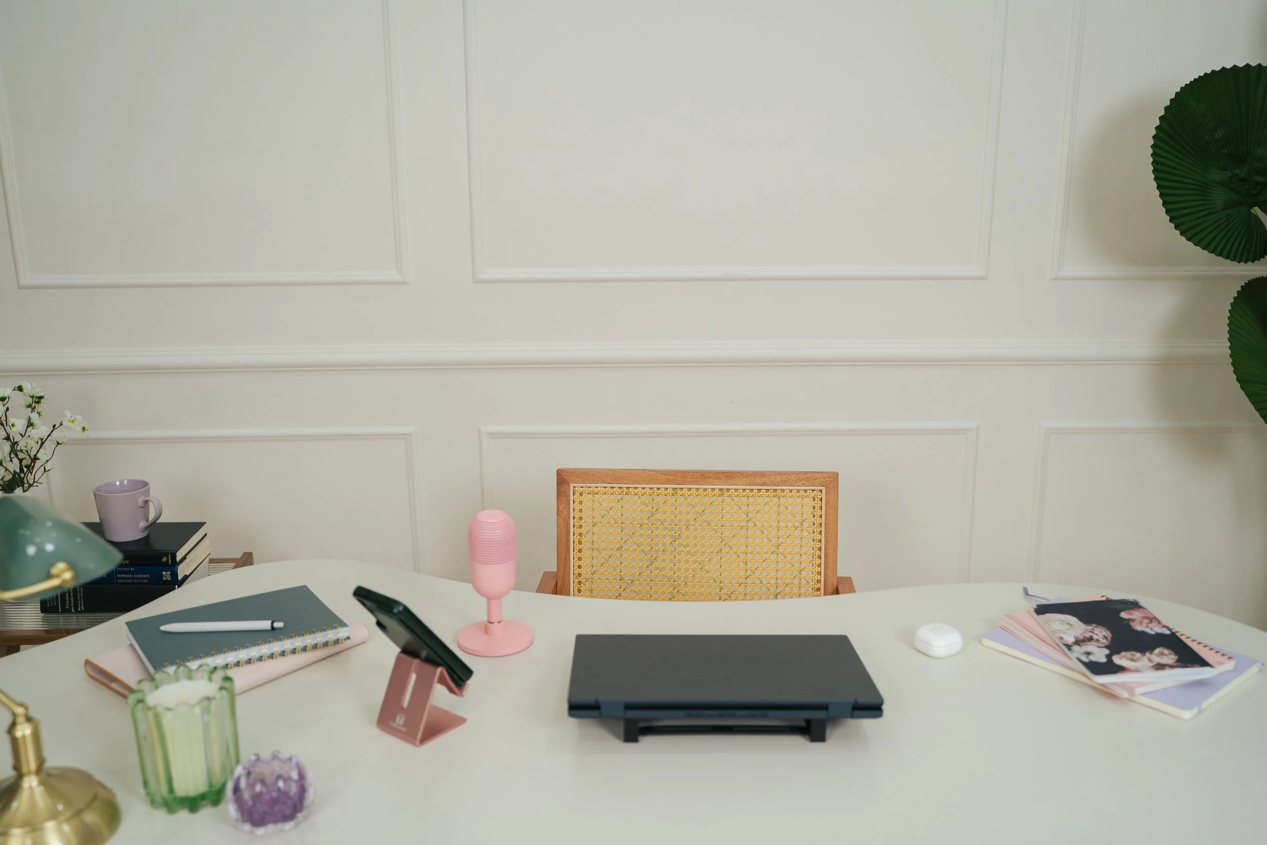 A modern workspace with a white desk, beige wall with paneling, and various office supplies including notebooks, a pen, a pink microphone, a laptop, a phone, and decorative items like candles, a plant, and a magazine. A wooden chair with a woven backrest is positioned at the desk.