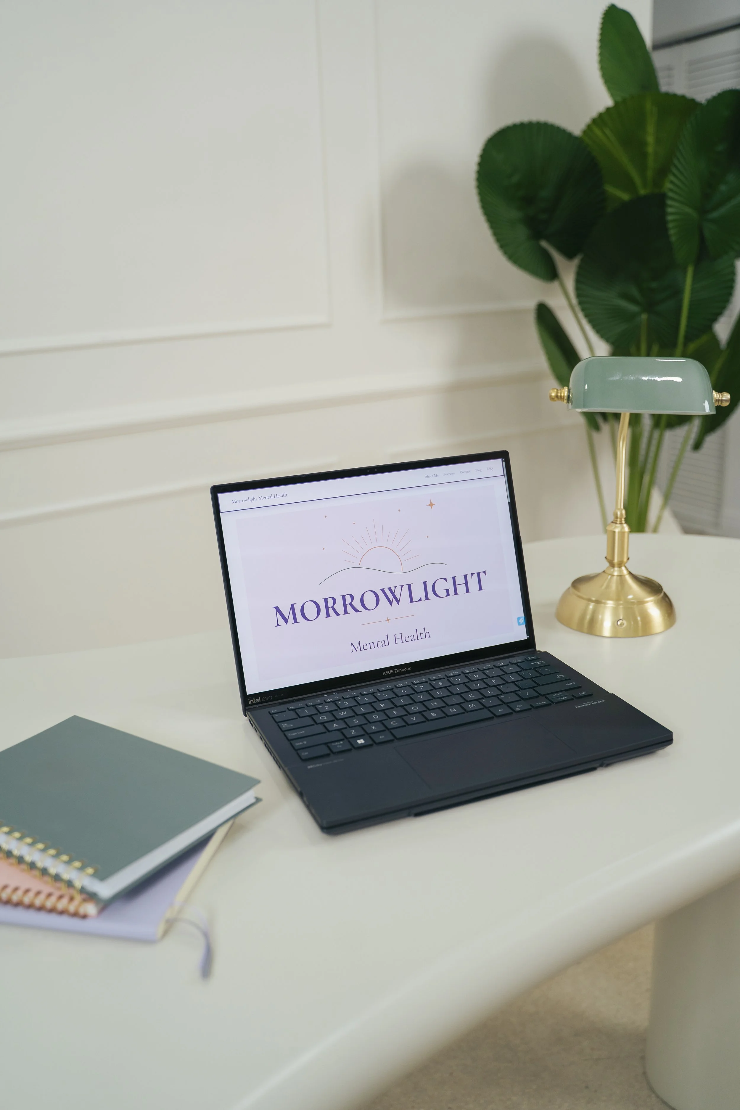 A workspace with a laptop displaying the logo for Morrowlight, a potted tropical plant, a green desk lamp with gold accents, and a closed notebook.