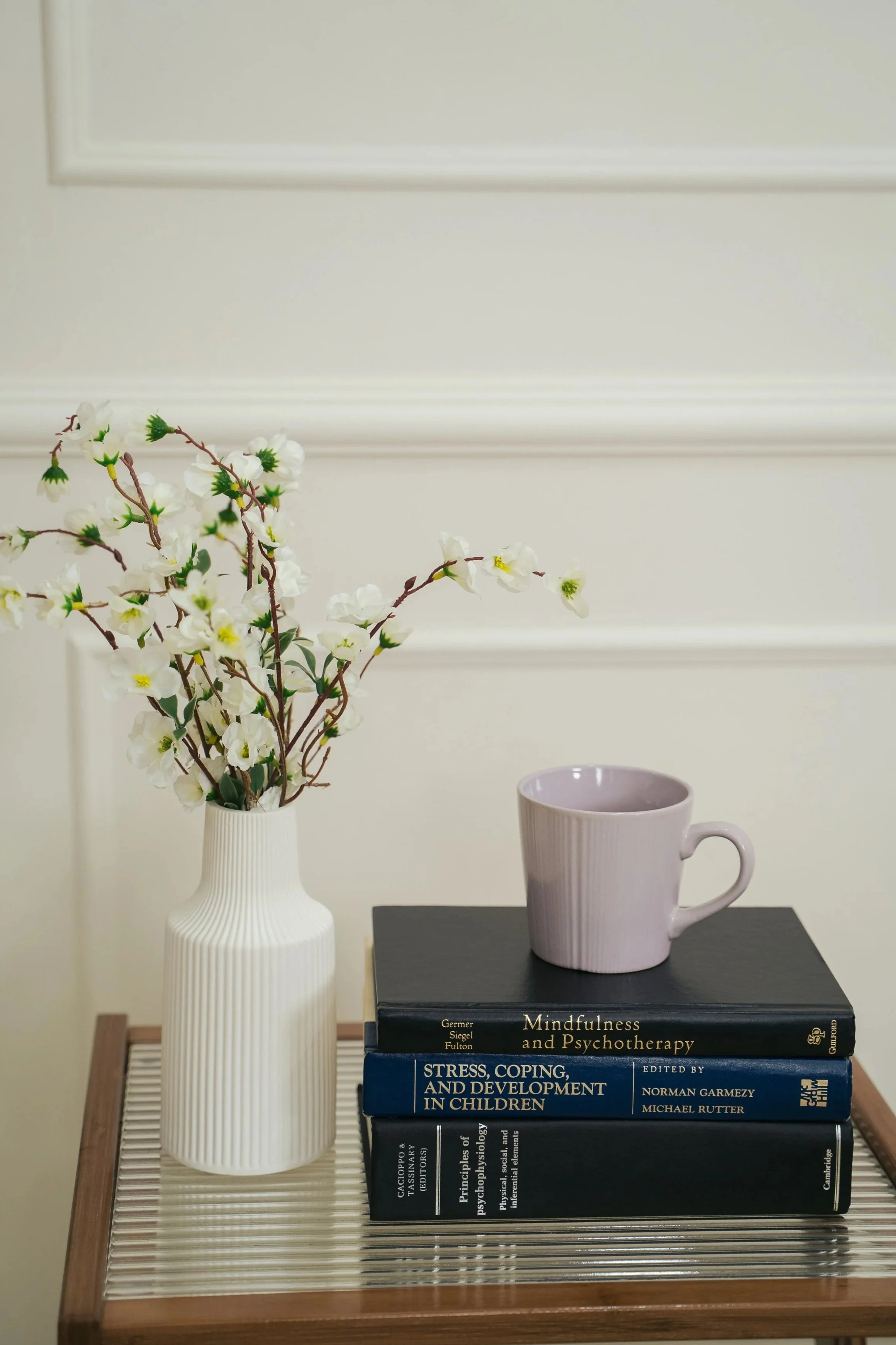 White vase with white flowers, stack of three books on psychology and child development, pink mug on top of the books, on a wooden table with a glass top, against a light-colored wall with decorative trim.