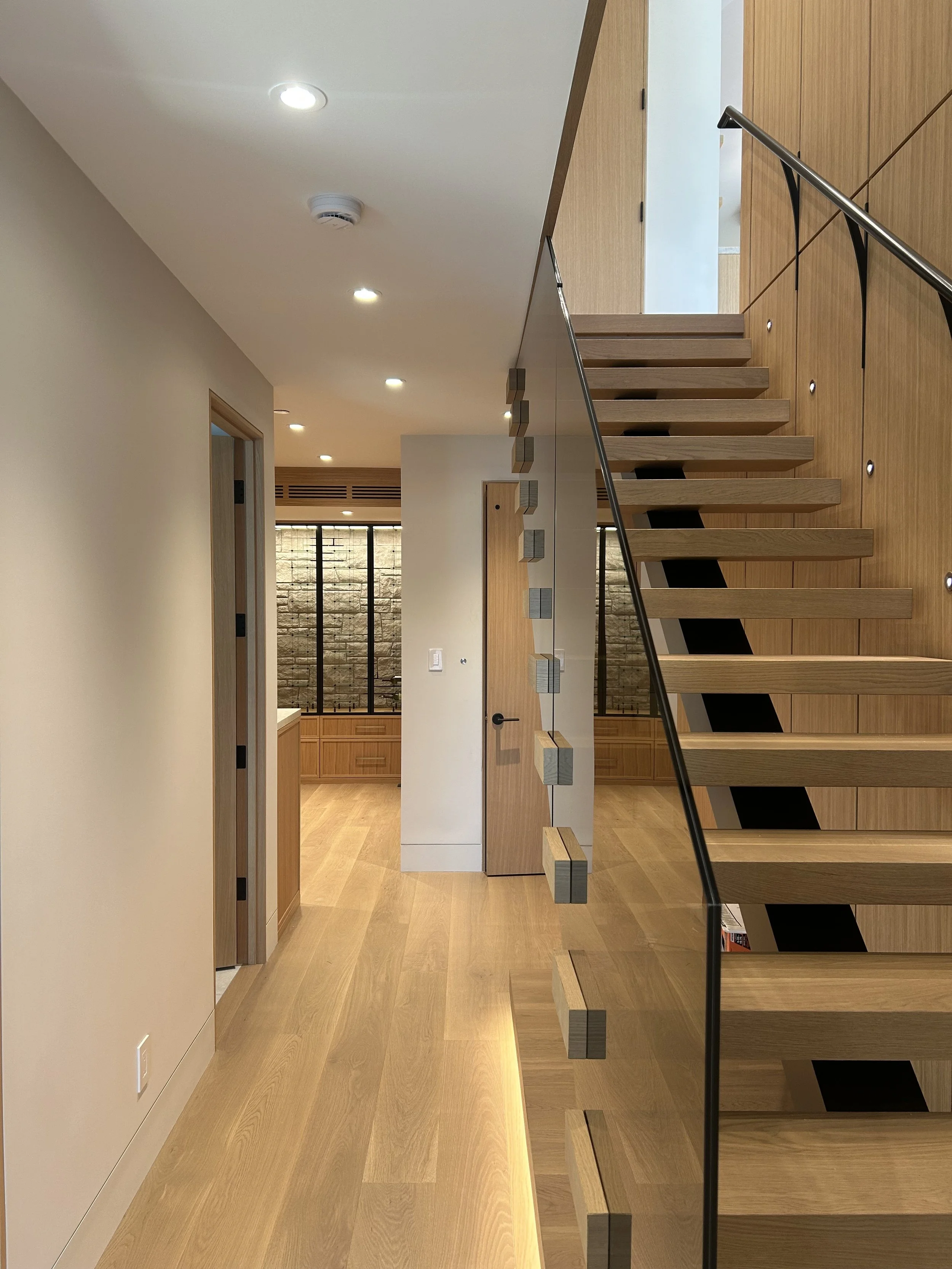 Interior view of a modern home with wooden stairs, a hallway with wooden flooring, recessed ceiling lights, and a wall with glass window panels.