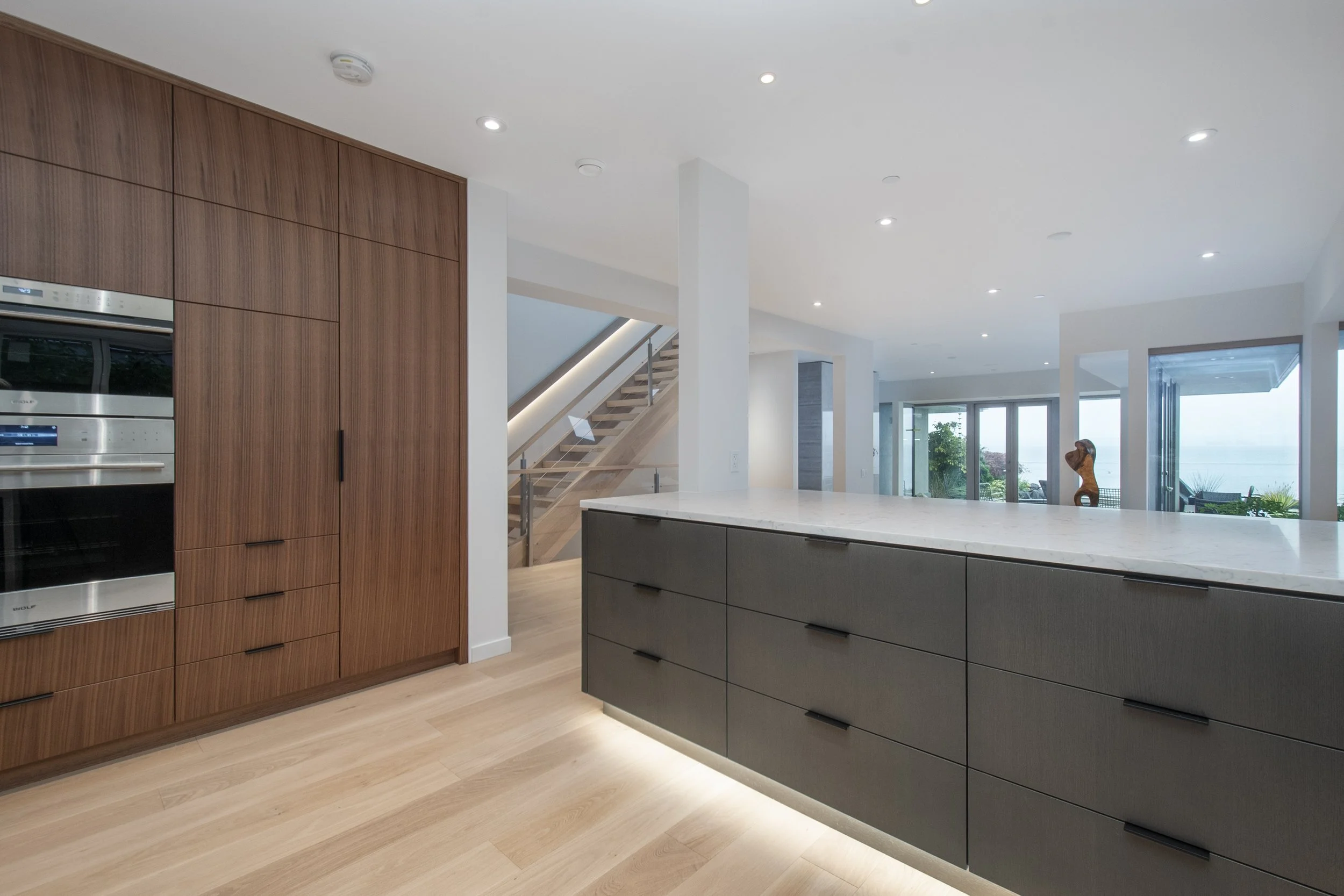 Modern open-concept kitchen with wooden and gray cabinetry, a large island with a white countertop, and large windows showing an outdoor view.