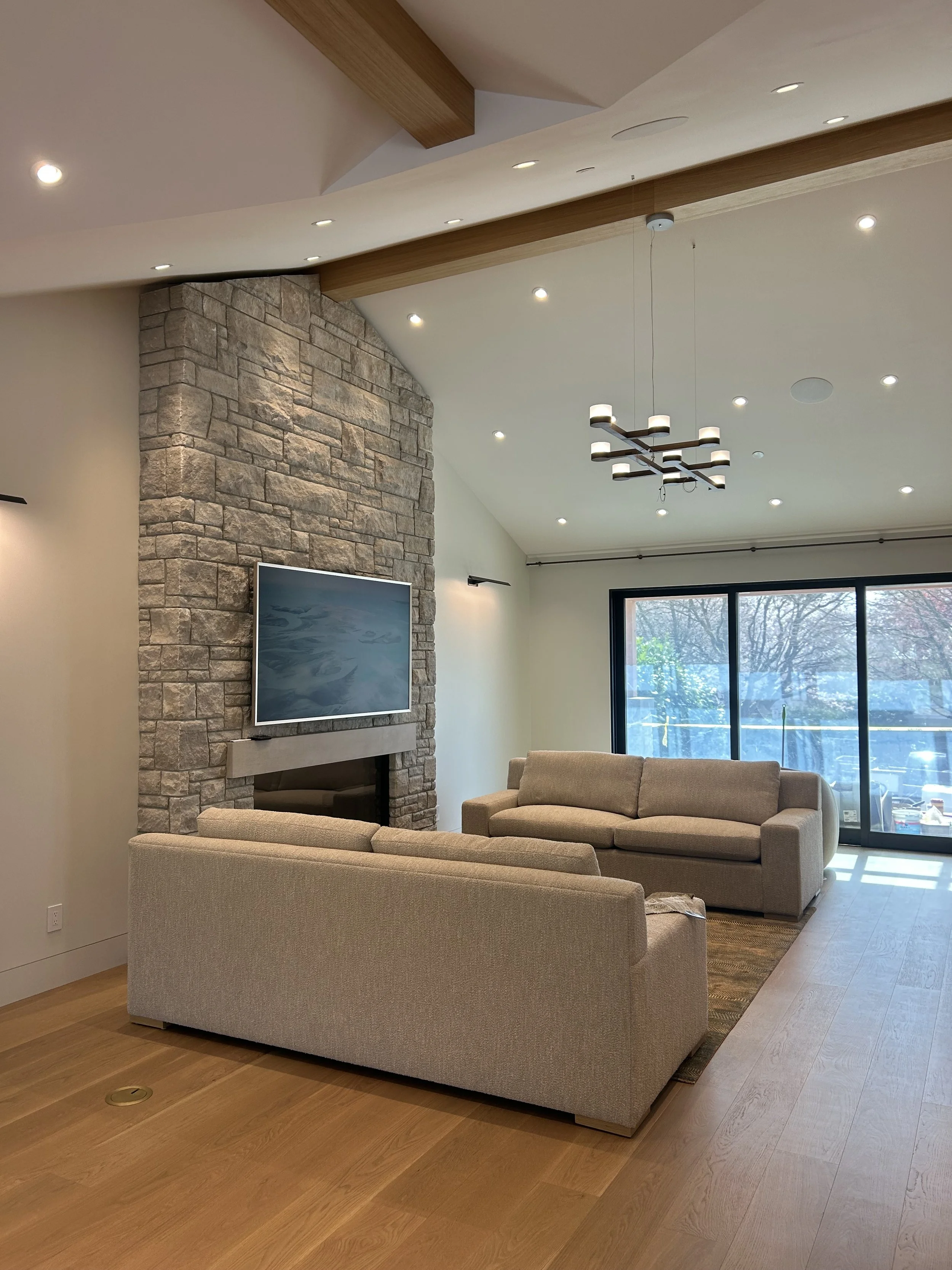 Interior living room with beige sofas, a stone fireplace with a mounted TV, large windows showing trees outside, and a modern chandelier hanging from the ceiling.