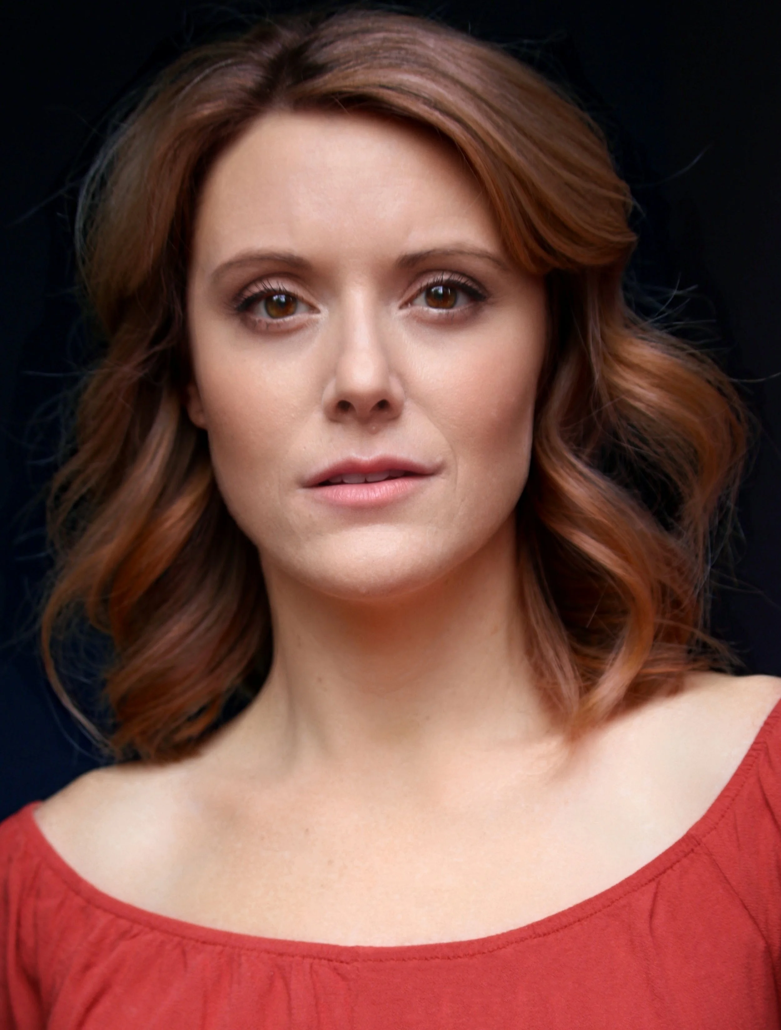 Close-up of a woman with red hair wearing a red top, standing against a dark background.