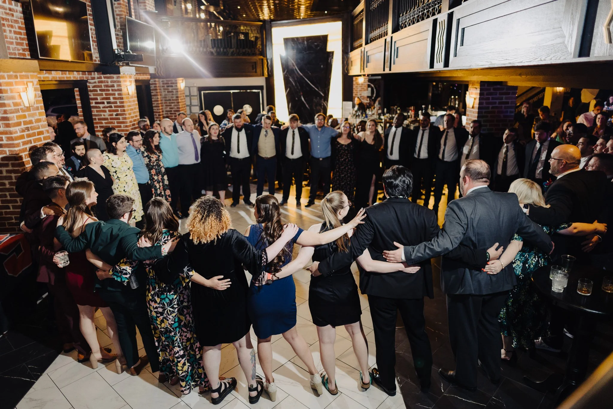 A group of people standing in a circle in a large ballroom sing with their arms wrapped around each other.