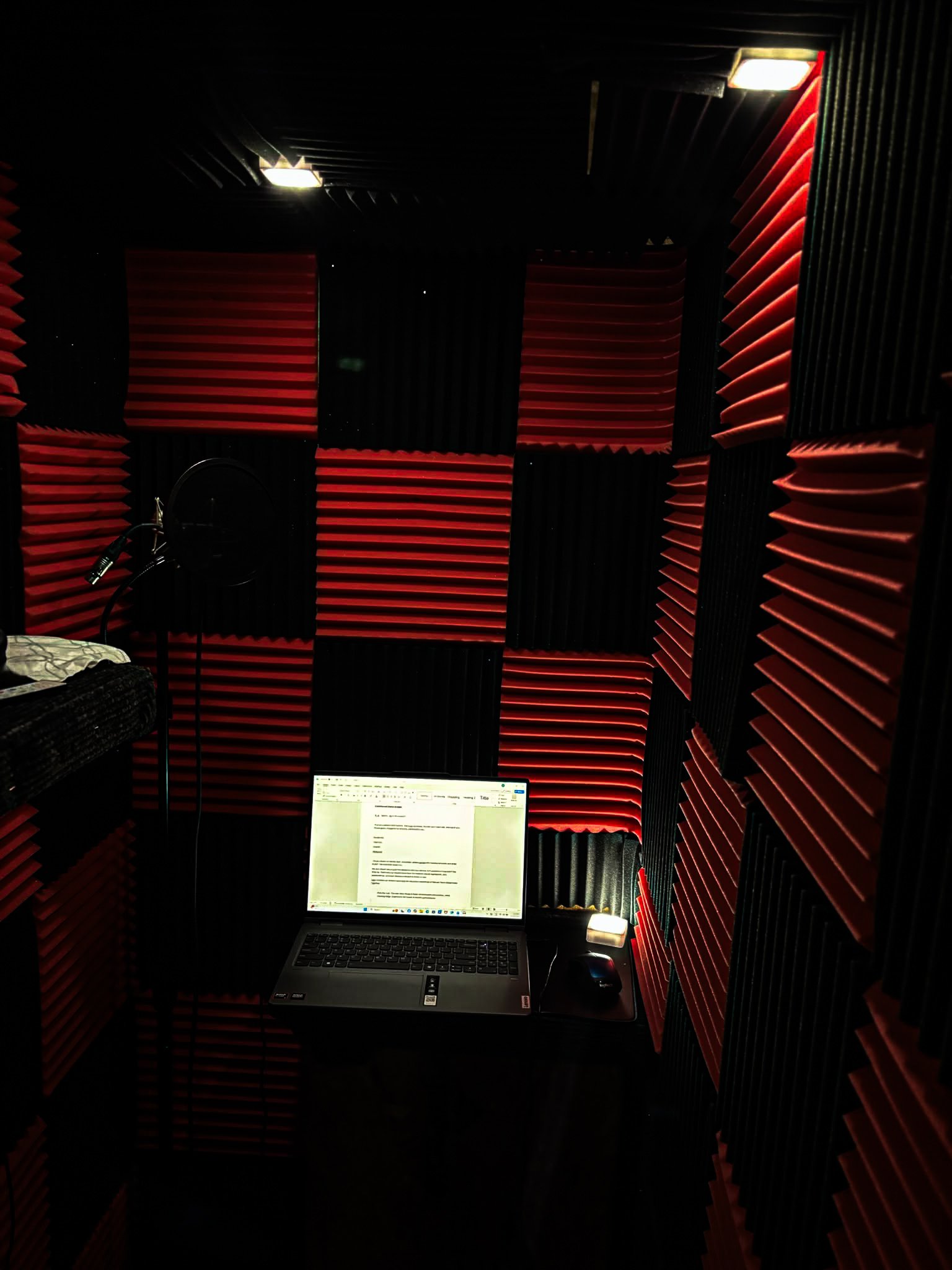 A recording booth with sound-absorbing red and black foam panels on the walls. A laptop displaying a document, a computer mouse, and a microphone are visible on a desk inside the booth.