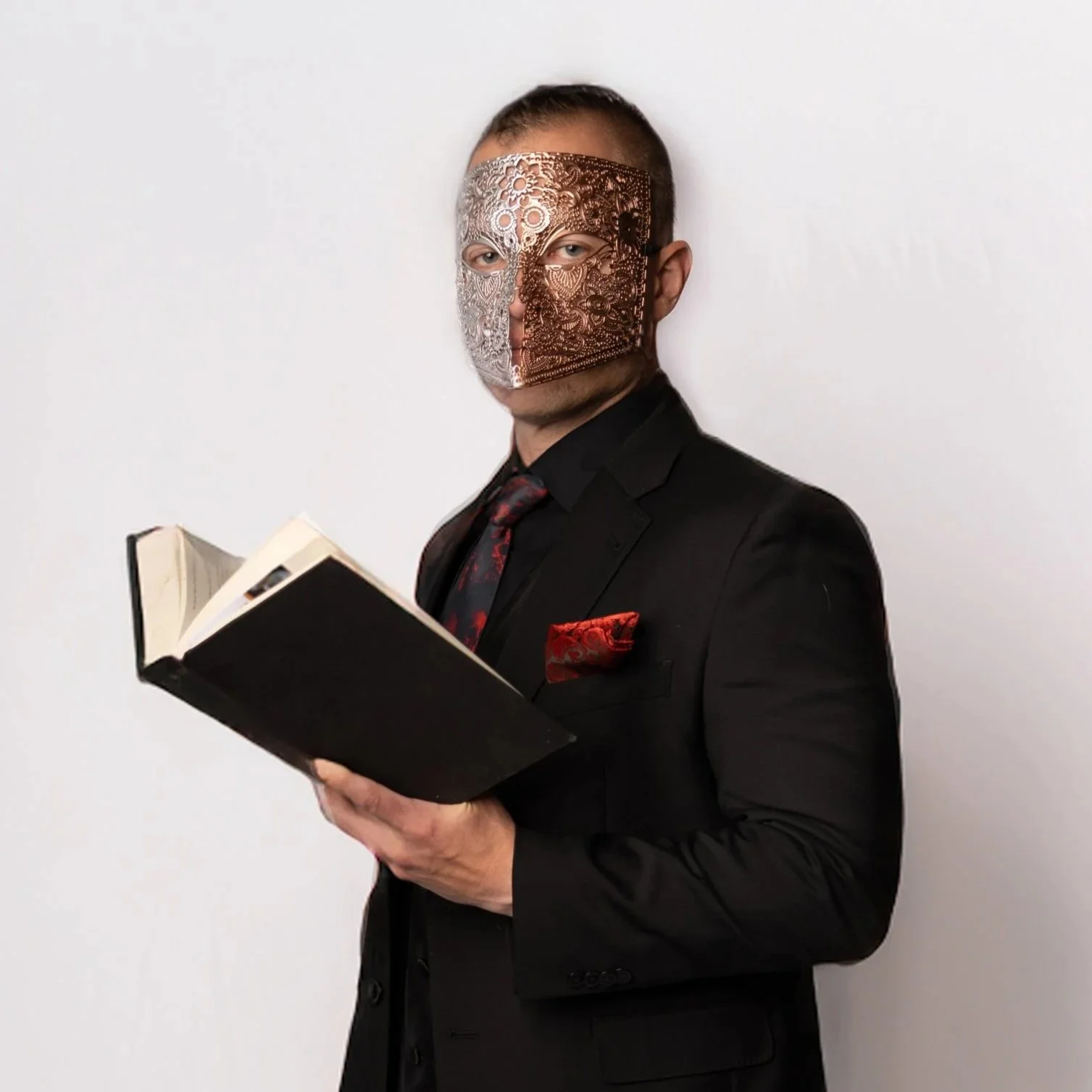 A man wearing a black suit and red tie with a red pocket square, holding an open book, and wearing a decorative half-metallic mask covering one side of his face, standing against a plain white wall.