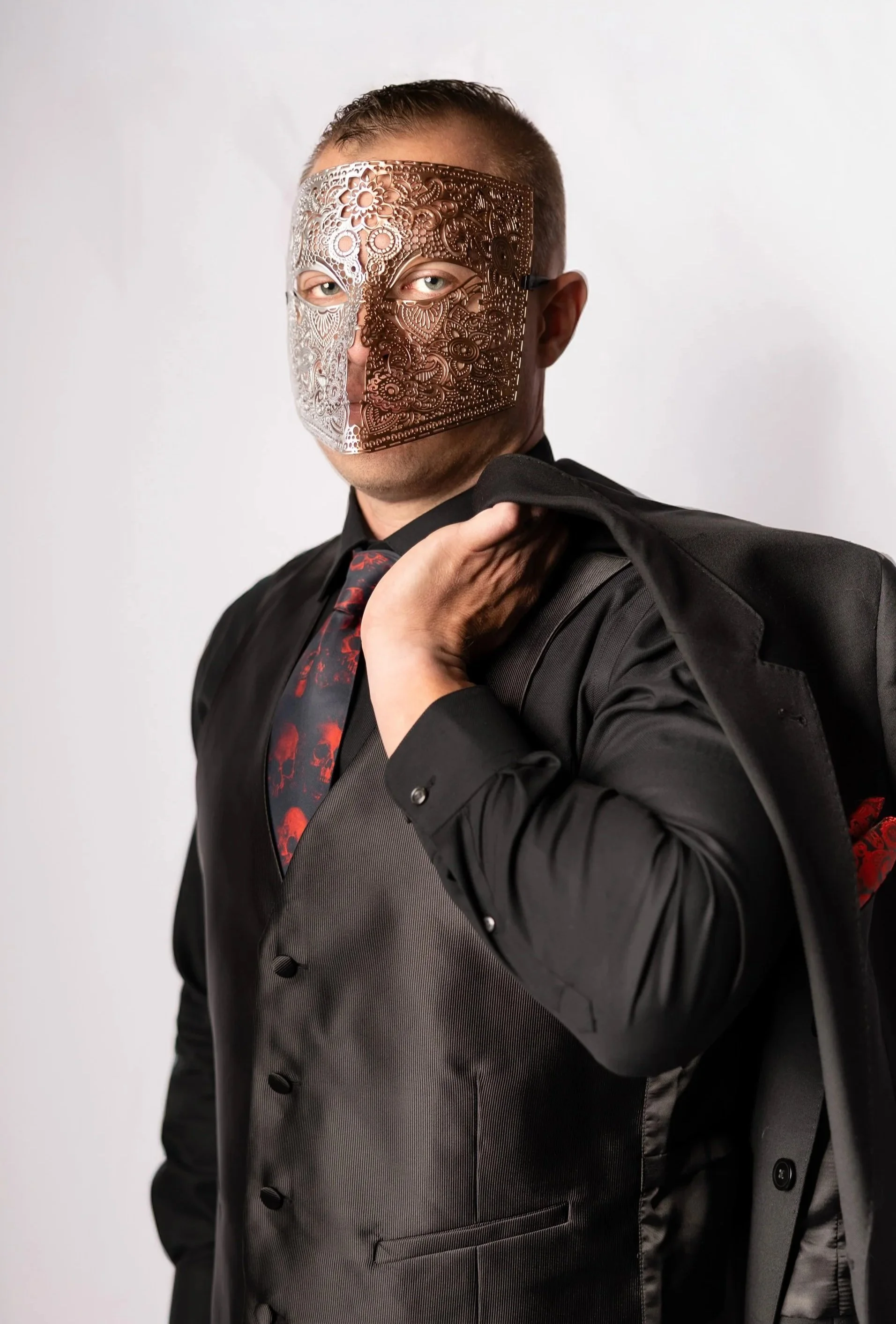 Man wearing a black suit with red patterned tie, striped vest, holding a gray coat over shoulder, and wearing a decorative metallic mask with floral and intricate pattern.