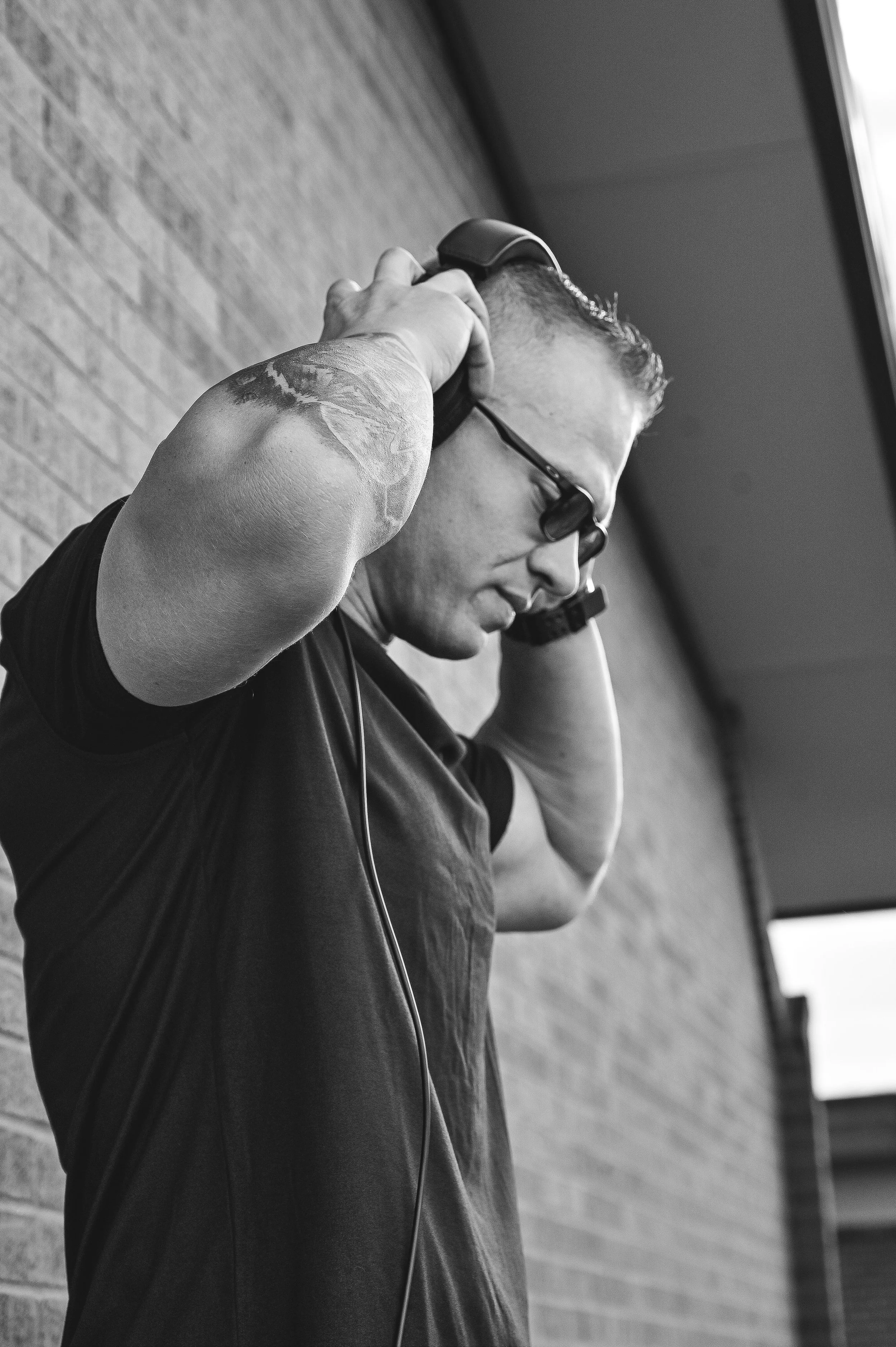 A man with glasses and tattoos on his left arm, wearing headphones and a black T-shirt, is adjusting or putting on his headphones indoors near a brick wall.