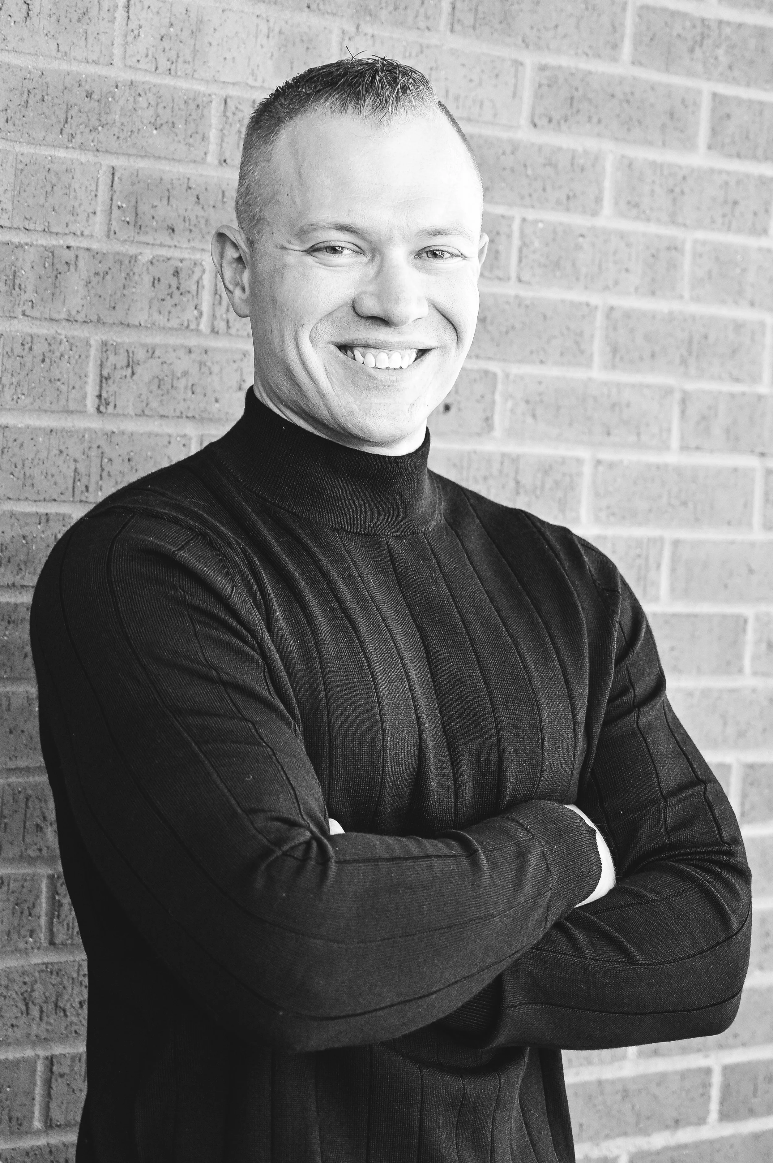 A smiling man with short hair wearing a dark ribbed sweater with arms crossed, standing against a brick wall.