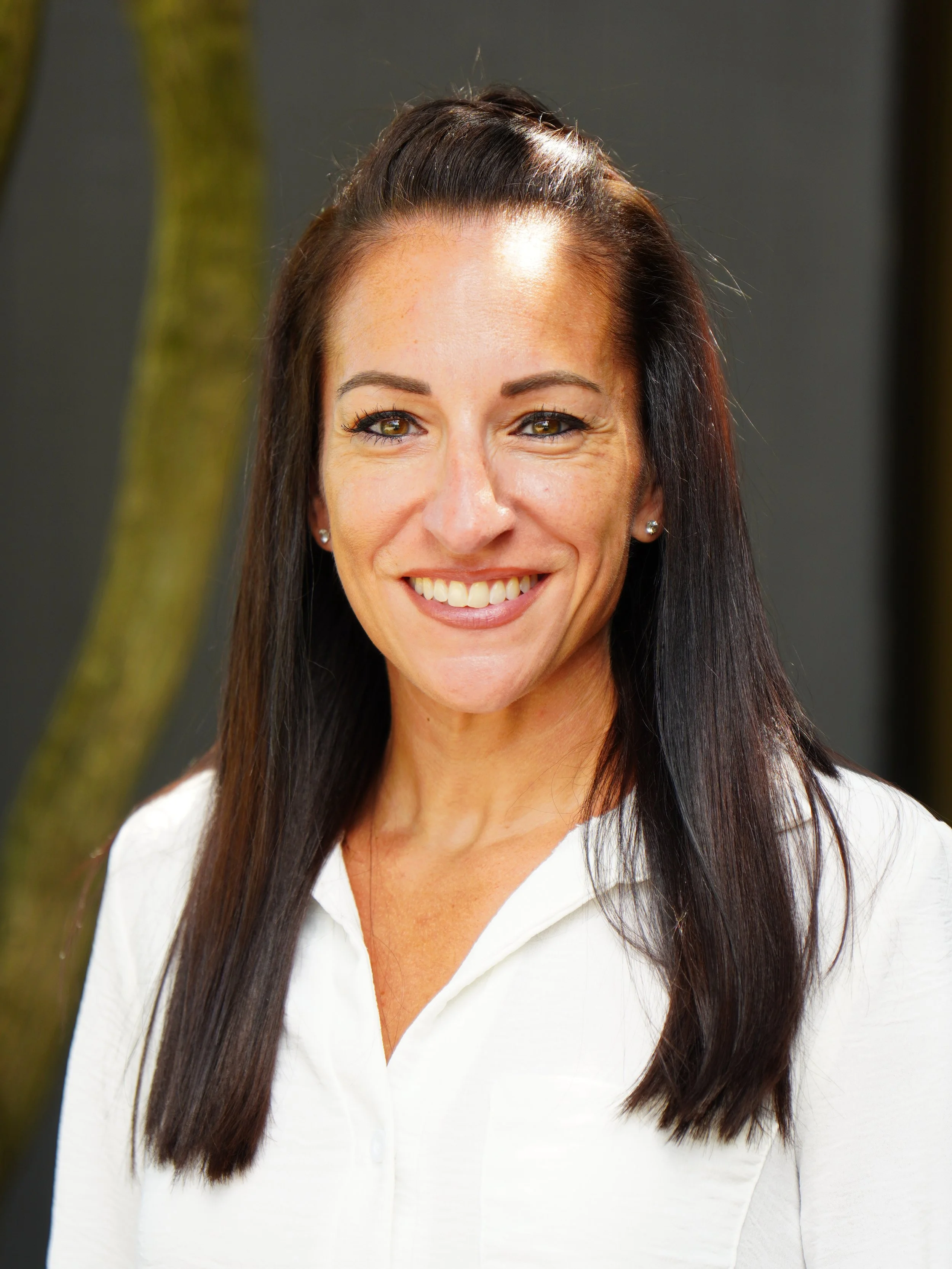 Close-up of a smiling woman with long dark hair, wearing a white shirt, standing outdoors in front of trees.
