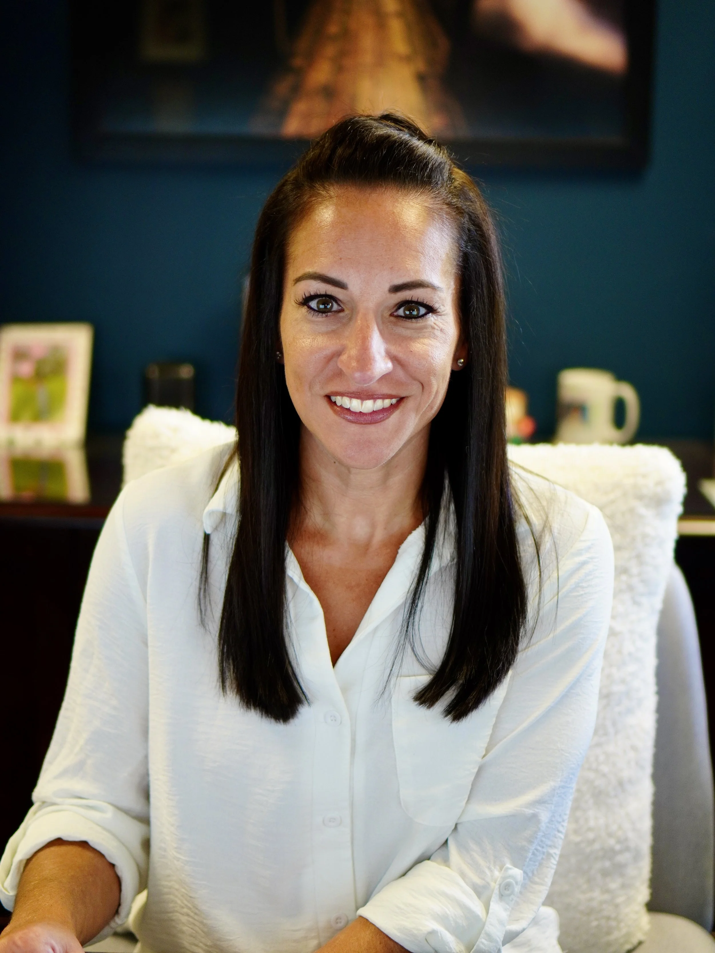 A woman with long dark hair, wearing a white shirt, smiling, sitting indoors with a dark blue wall, framed pictures, and a mug in the background.