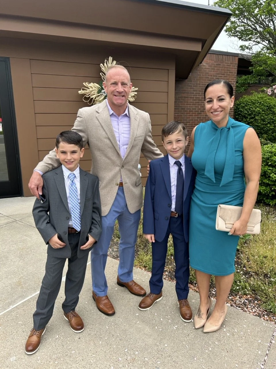 A family of four dressed formally stands outside a building, smiling at the camera. The man wears a beige blazer and blue pants, the woman wears a teal dress and carries a beige clutch, and two boys wear suits with ties.
