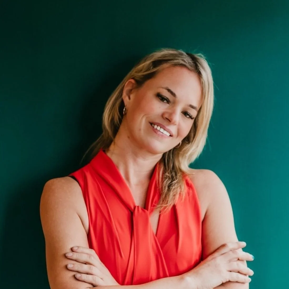 Person with blonde hair in a red sleeveless top against a green background, smiling with arms crossed.