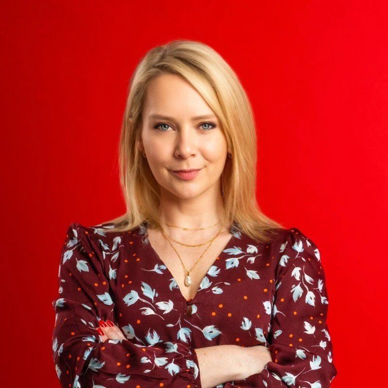 Person with blonde hair smiling, wearing a floral blouse, standing against a red background.
