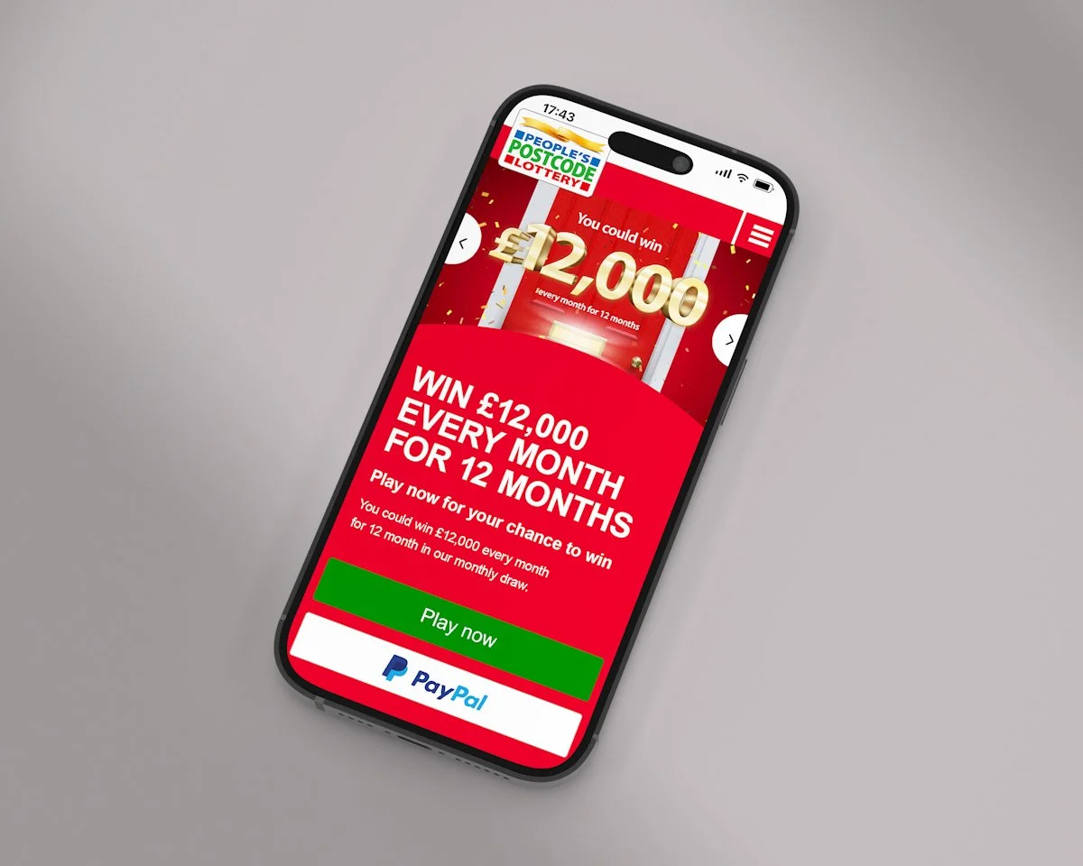 A smartphone displaying a lottery promotion for People's Postcode Lottery with the text 'Win £12,000 every month for 12 months' and a green 'Play now' button.