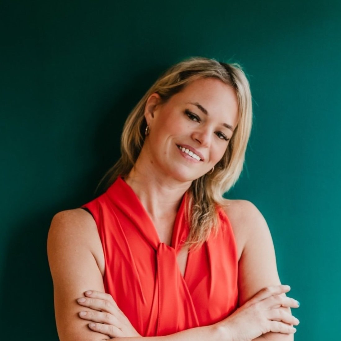 Person with blonde hair in a red sleeveless top against a green background, smiling with arms crossed.