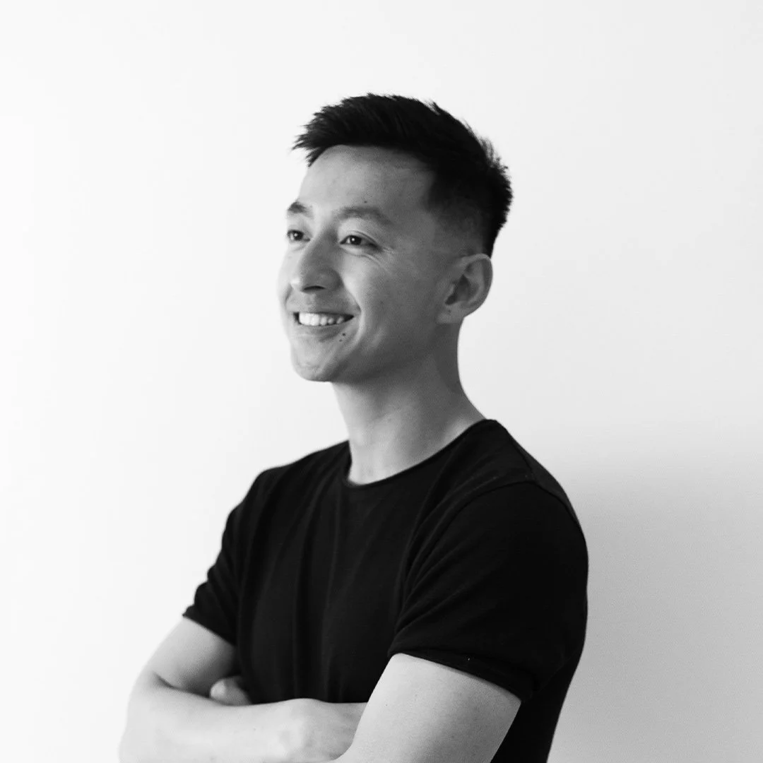 Black and white photo of a smiling man with arms crossed, wearing a black t-shirt, standing against a plain light background.