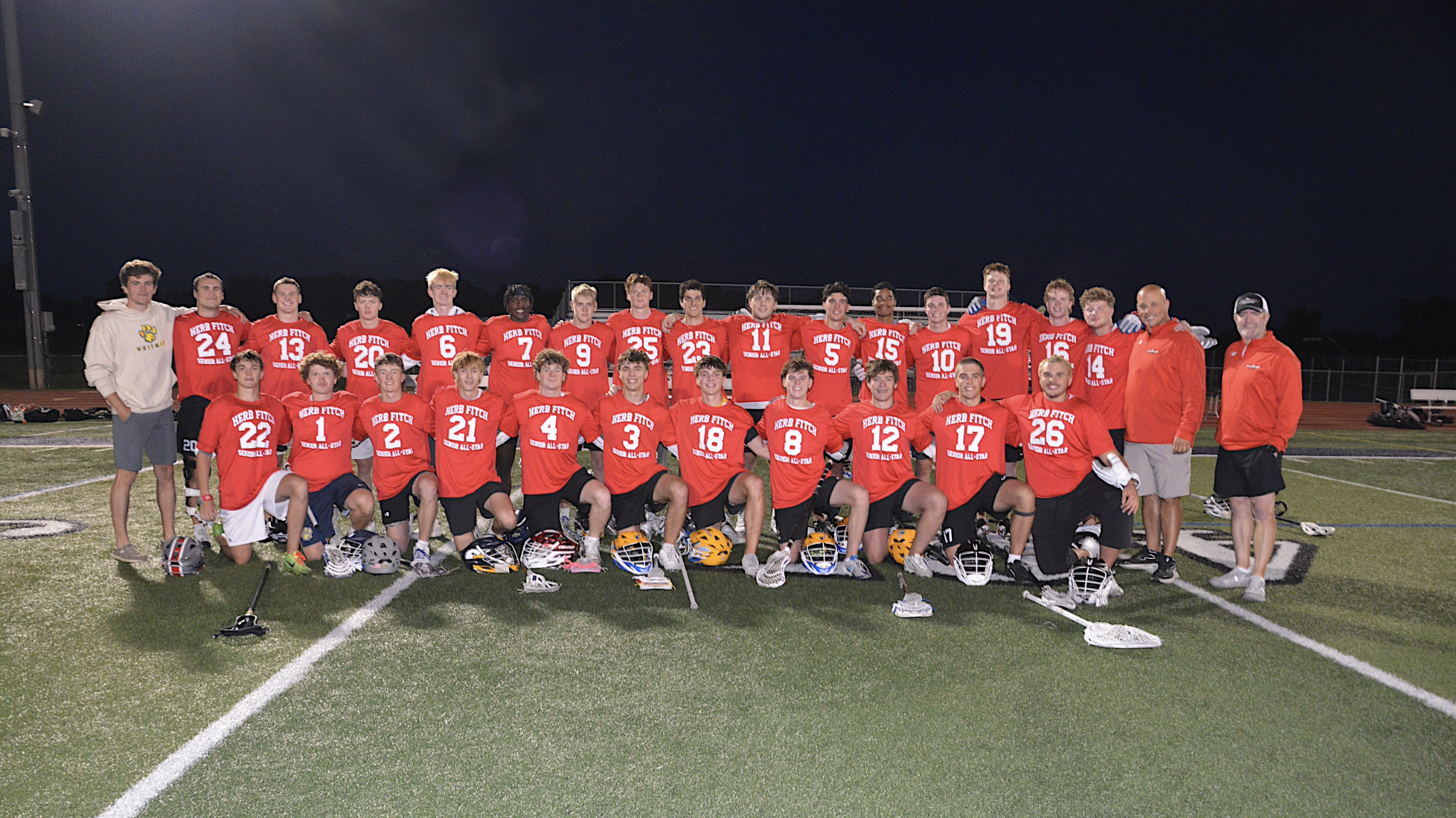 Group of young male football players and coaches standing on a football field at night after a game or practice. The players are dressed in red jerseys and black shorts, with helmets on the ground in front of them. Some players are kneeling in the front row, and others are standing behind. Two coaches are on the right, wearing red jackets, and a young man in a hoodie and shorts is on the left. The background is dark with stadium lights illuminating the scene.