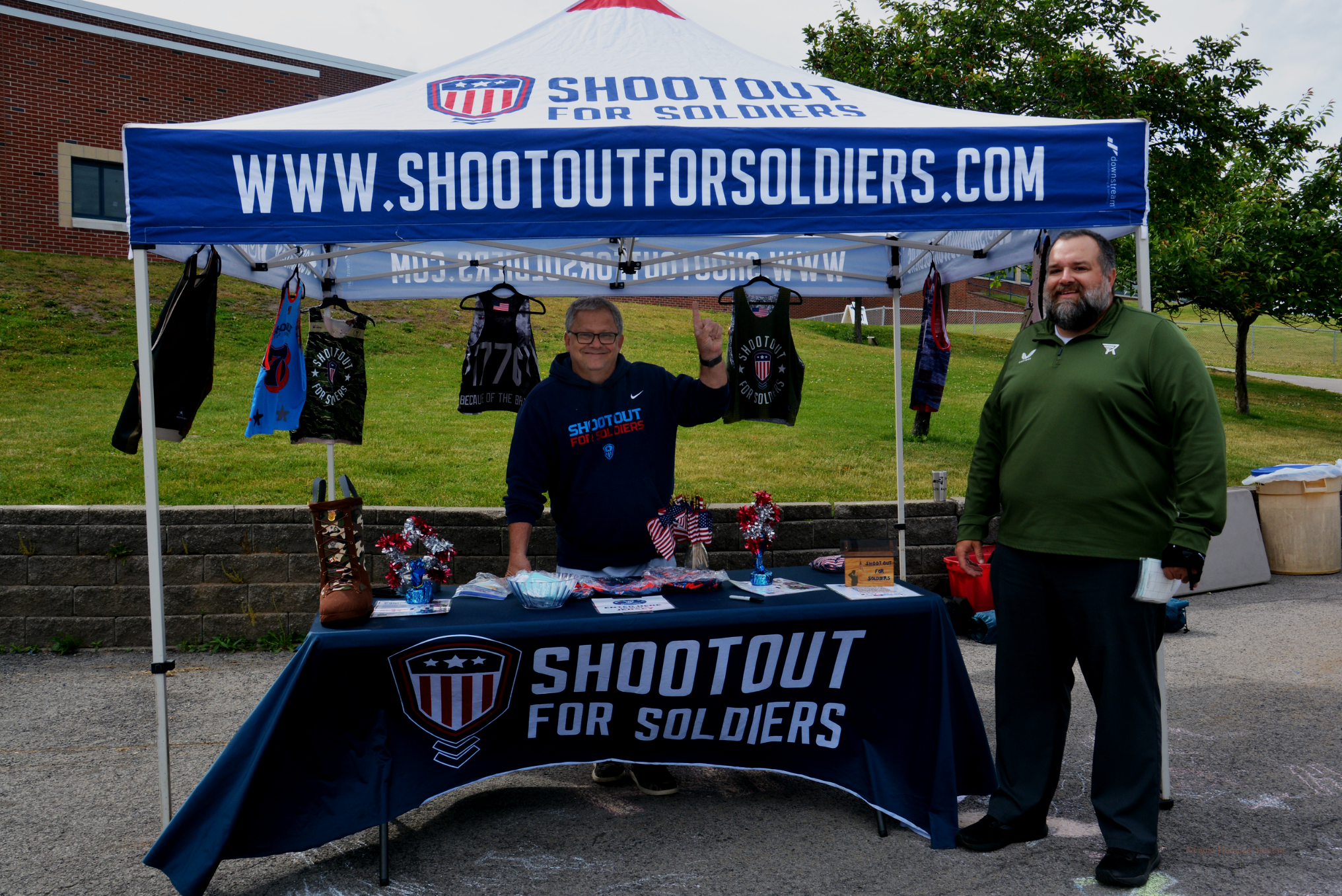 A booth with a blue canopy that reads 'Shootout for Soldiers' and a website URL 'www.shootoutforsoldiers.com.' Two men are standing in front of the booth, smiling. One is wearing glasses and a navy hoodie with the event's logo, and the other has a beard and is wearing a green shirt. The booth displays various shirts, decorations, and promotional items on a table.