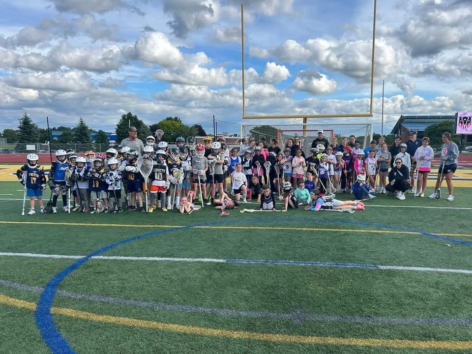 A large group of children and adults gathered on a sports field for a group photo, many of whom are wearing sports gear and helmets, with a goalpost and some equipment visible in the background.