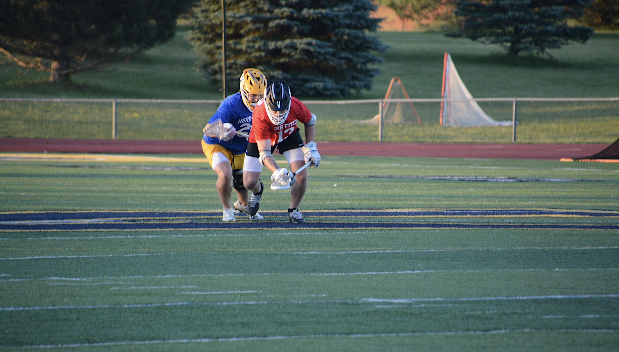 Two lacrosse players in protective helmets, gloves, and uniforms are on a green field with a track; one in red is bent over controlling the ball with a lacrosse stick, while the other in blue is close behind.