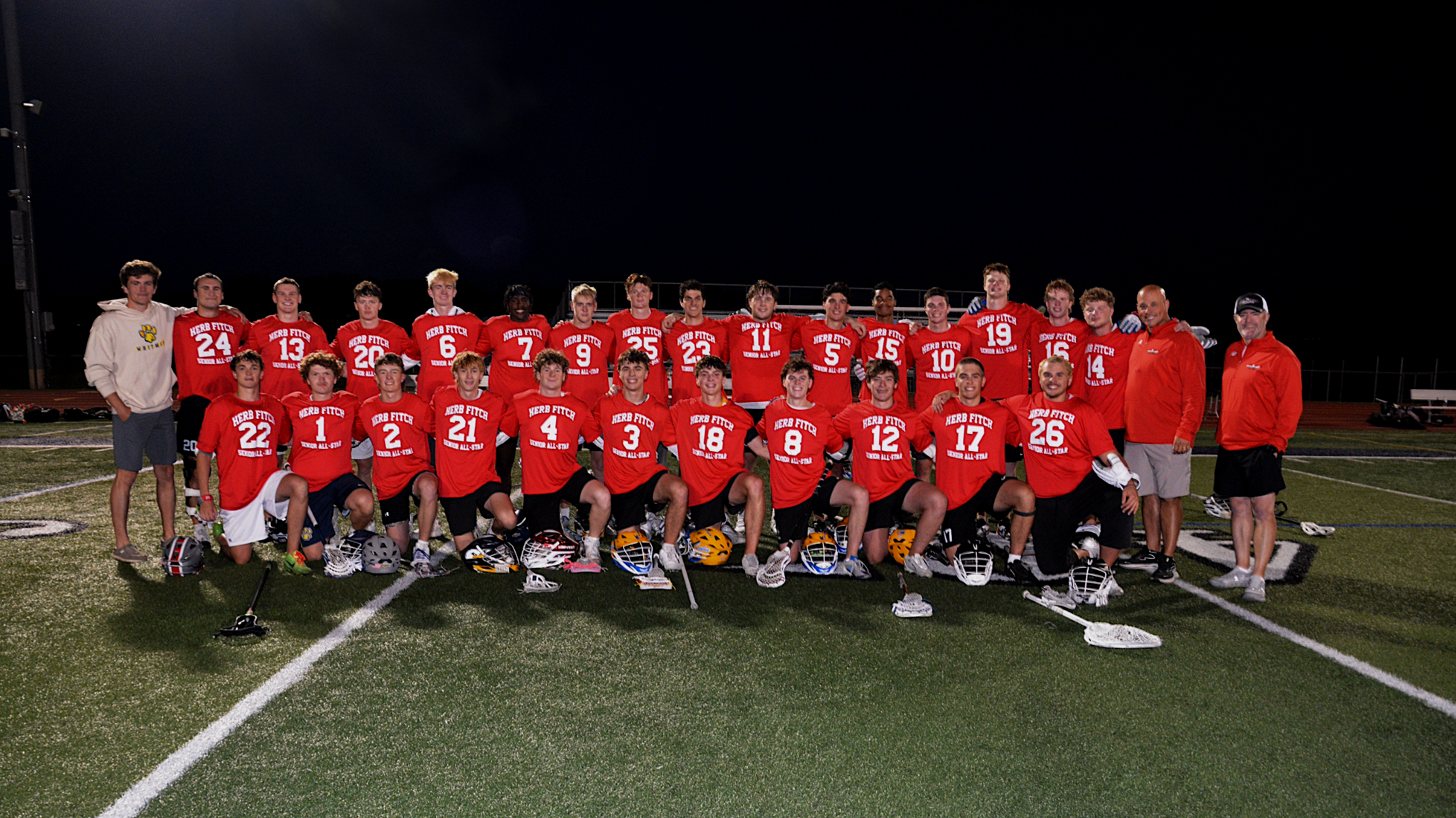 Group of young male football players and coaches posing on a football field at night, wearing red jerseys with white lettering, and helmets placed in front of them.