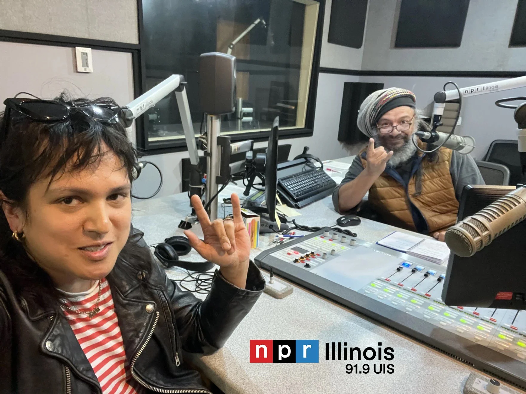 Two individuals in a radio studio with professional microphones, sound equipment, and a mixing console, both smiling and making hand gestures. One woman with glasses on her head and a leather jacket is in the foreground, while a man with a beanie and glasses sits in the background. The studio has soundproofing panels and branding for NPR Illinois 91.9 UIS.
