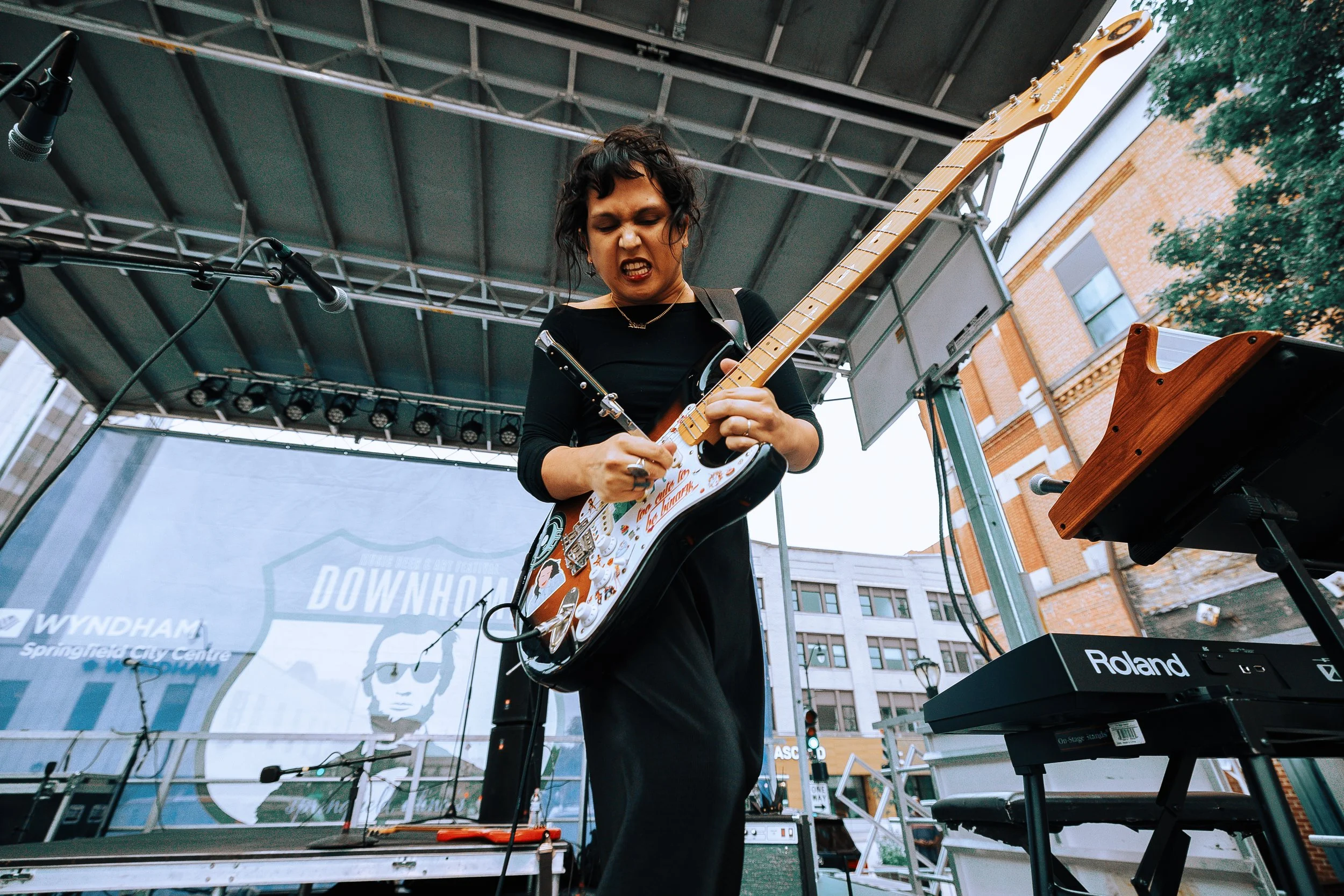 A female musician passionately plays an electric guitar on an outdoor stage with microphones and keyboard equipment. She is wearing a black top and has short, wavy dark hair. The stage is set against buildings and a tree is partially visible on the r