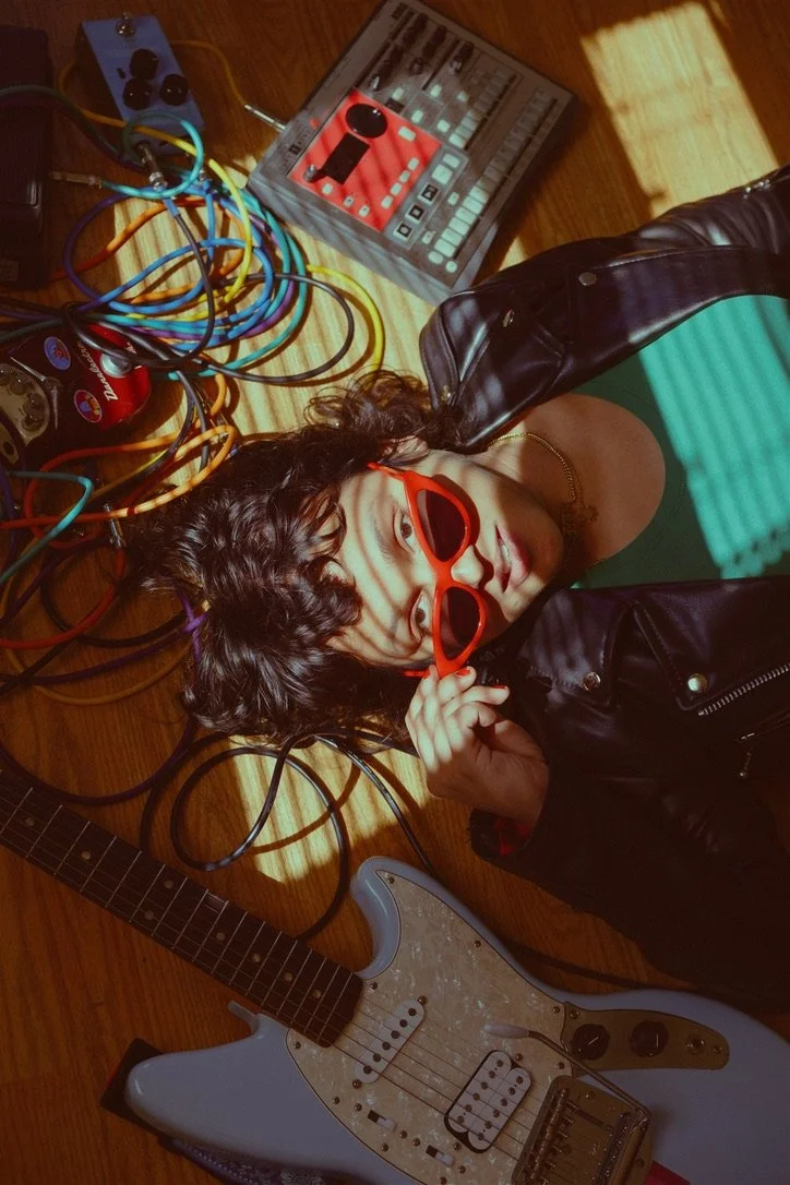 A person lying on a wooden floor surrounded by musical equipment, including a guitar, a mixer, a pedal, and multiple cables. The person is holding red sunglasses and wearing a black leather jacket and a green top, with shadows from blinds cast across their face.
