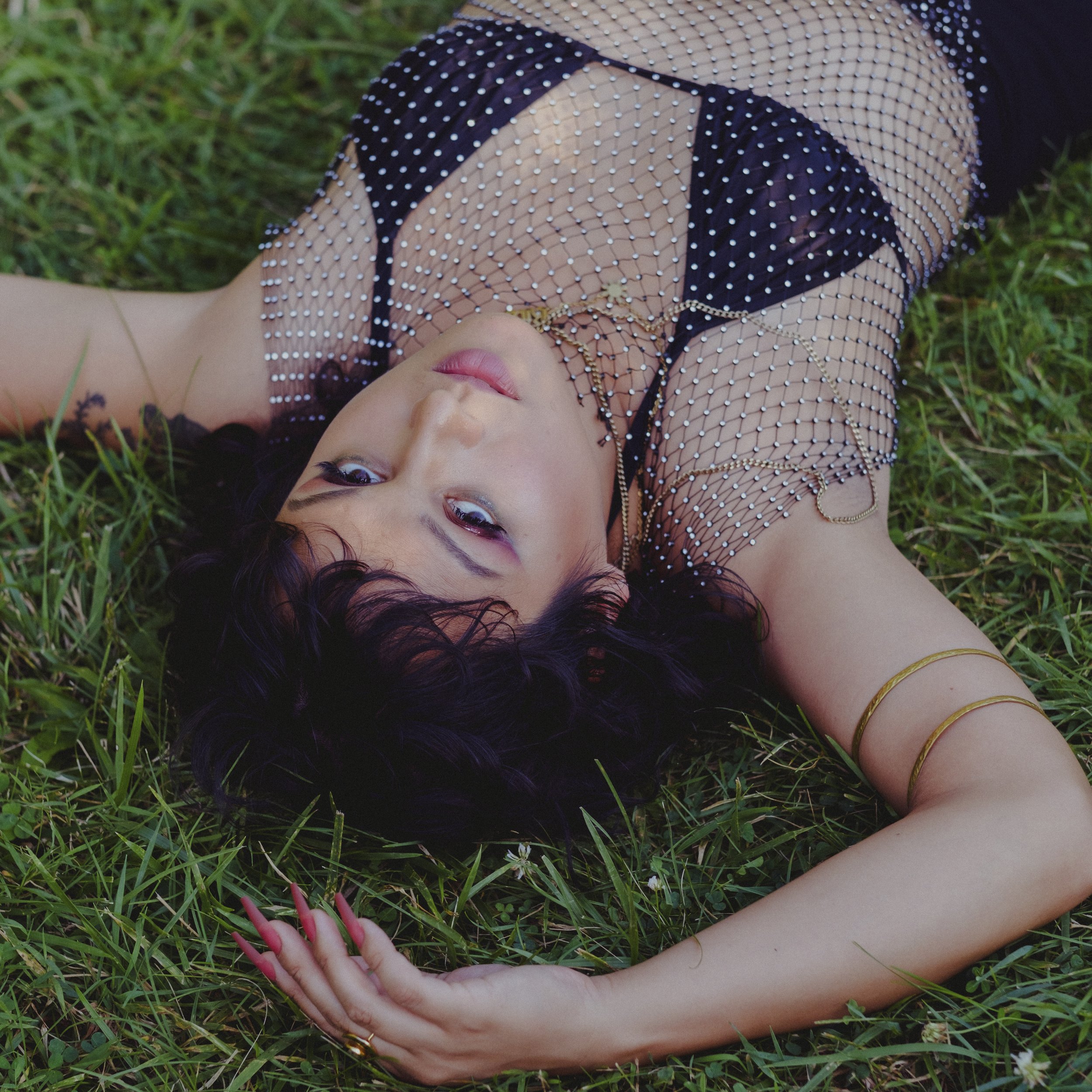 A woman lying on her back on green grass, wearing a black mesh top with white rhinestones and gold jewelry, including rings and bracelets; her hand with pink nails is visible in the foreground.