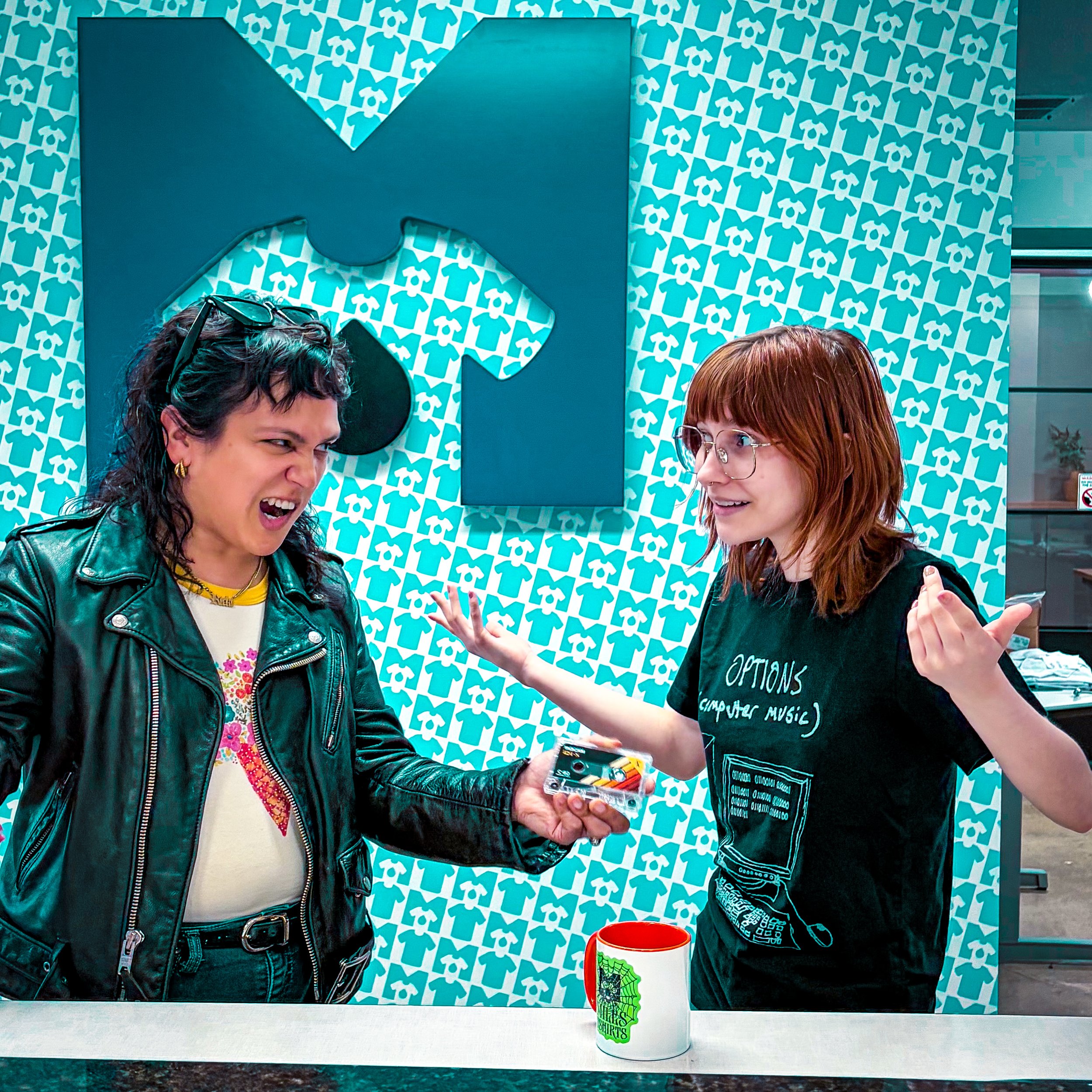 Two women with short hair, one with glasses, are engaged in conversation at a table with a nearby wall decorated with a pattern of blue t-shirts and dog faces. A white cup with a green and black design and a red rim is on the table.