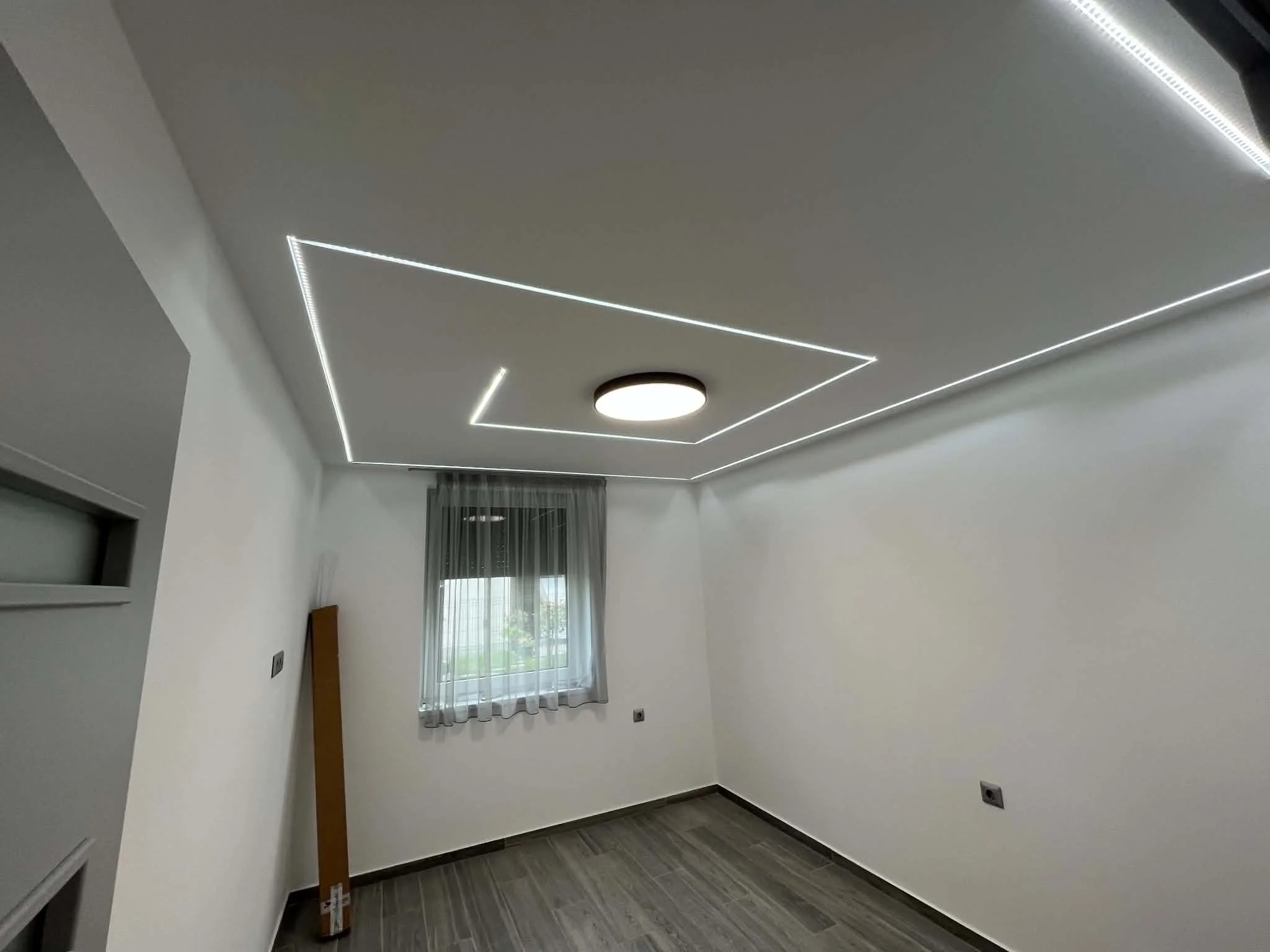 Empty room with white walls, a window with sheer curtains, a ceiling light fixture, and LED strip lighting on the ceiling.
