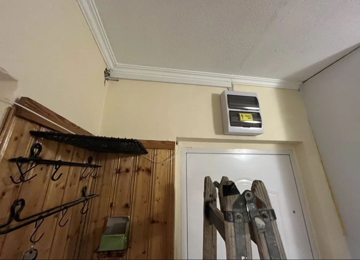 Interior of a room with a wooden wall, metal hooks, a small shelf, a ladder, and an electrical breaker box on the wall.