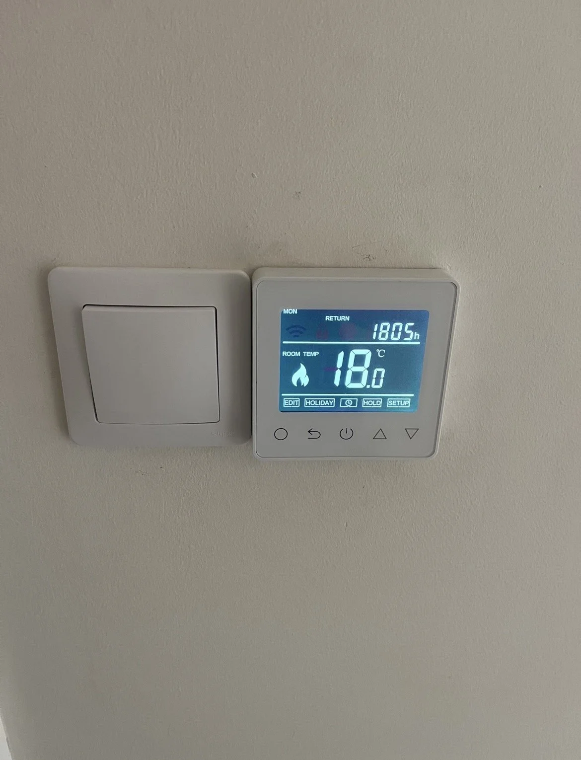 Digital thermostat on a wall showing room temperature of 18.0°C and 1805 hours time display, next to a blank white switch cover plate.