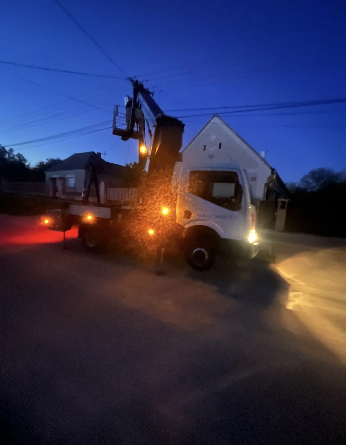 A utility truck is parked on a residential street at dusk, with its bucket lift extended to a utility pole for maintenance. The truck has orange safety lights and the street is dimly lit.