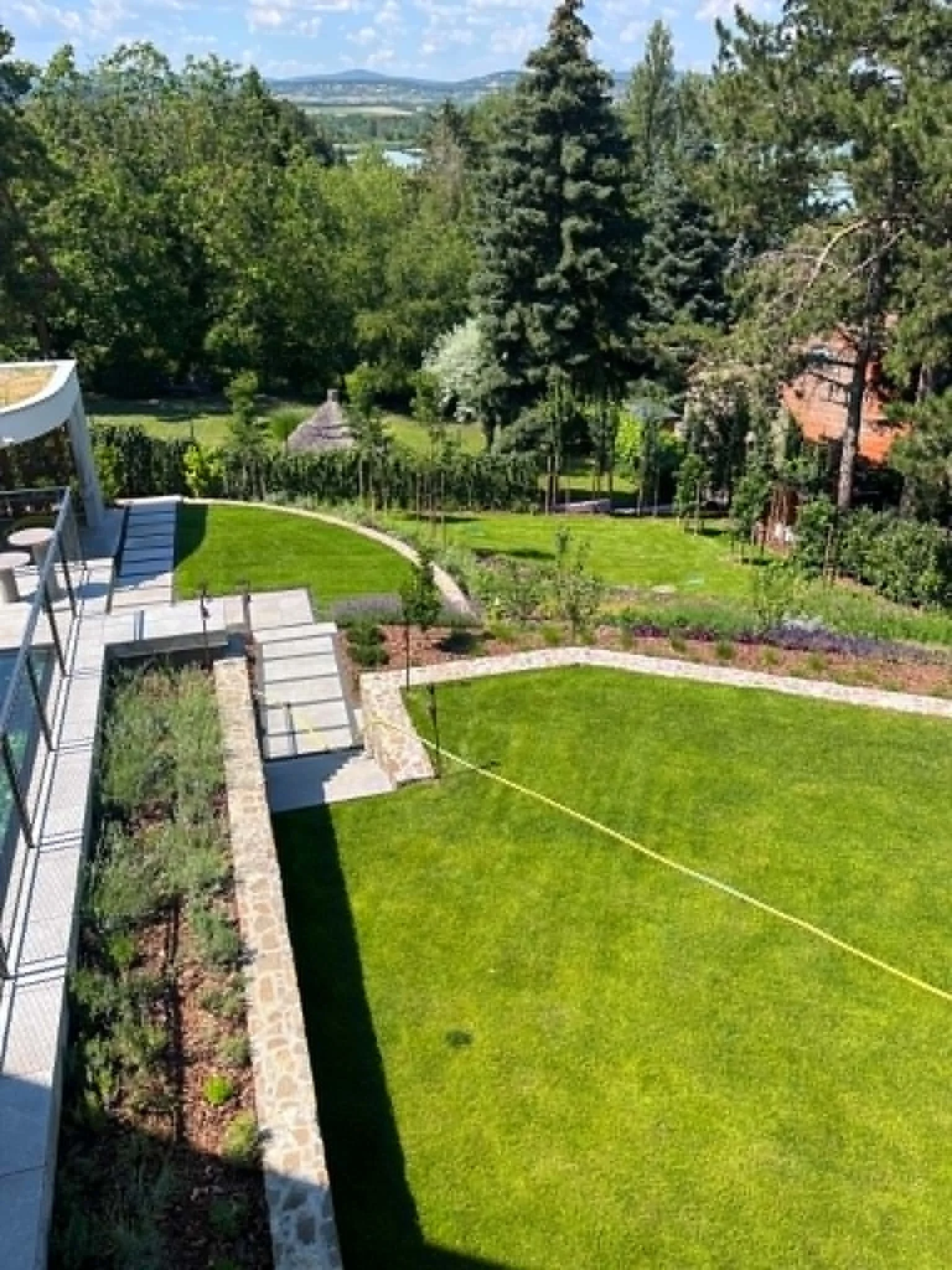A backyard garden with neatly trimmed bright green grass, stone pathways, trees, and shrubs, with mountains in the distance under a partly cloudy sky.