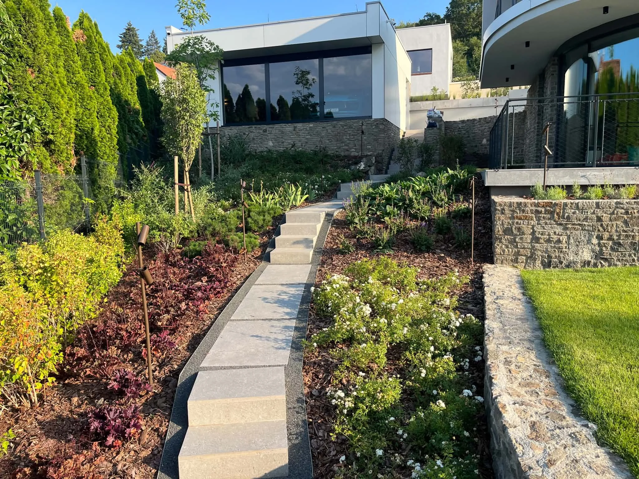 Modern house with large glass windows and a flat roof, surrounded by a landscaped garden with a stone pathway, green hedges, and flowering plants.