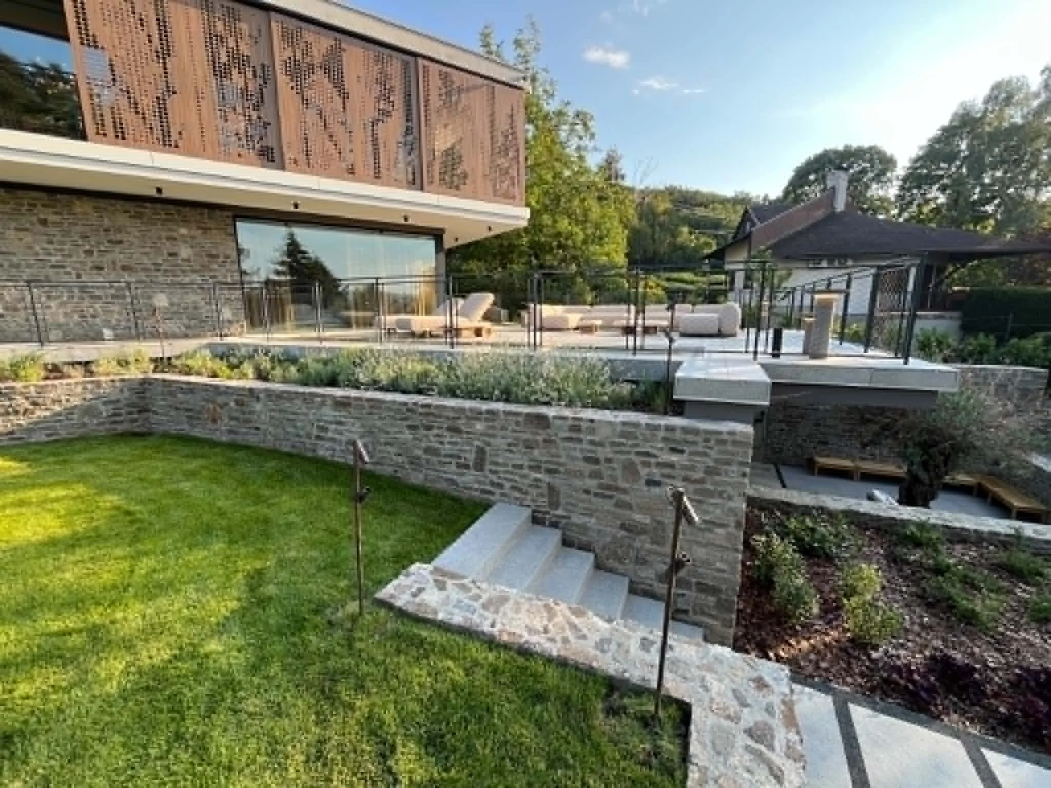 Exterior backyard view of a modern house with a stone wall, glass sliding doors, a small patio with lounge chairs, a gazebo, and a well-manicured lawn, surrounded by trees and greenery.