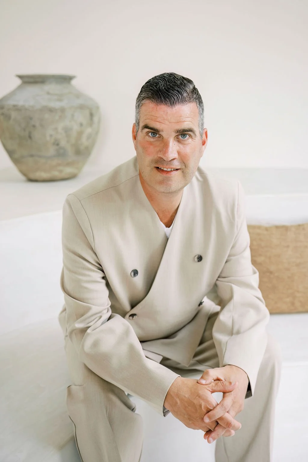 A man with dark hair and blue eyes wearing a beige chef's coat and pants, sitting on a white surface, with a large decorative urn in the background.