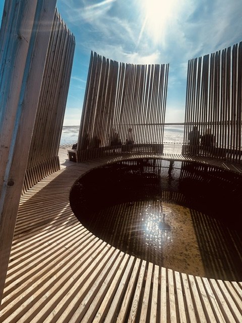 View of a beach scene taken through a wooden circular structure with vertical slats, with the ocean and sky visible in the background.